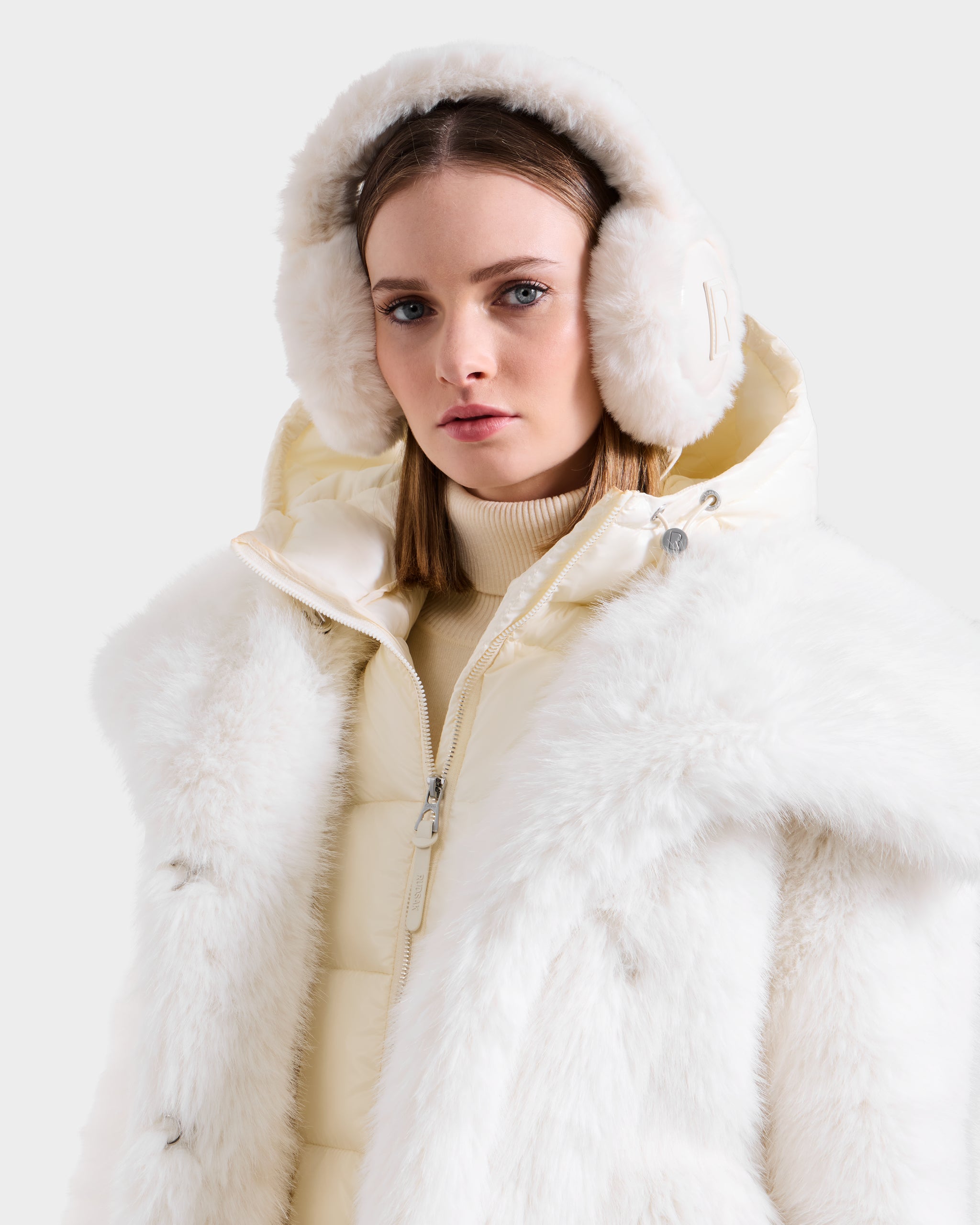 A woman wearing Rudsak’s VALENCIA - 8425543 Iceland earmuffs, a light cream turtleneck, and a white fur coat over a quilted jacket stands against a plain background, highlighting this chic winter accessory.