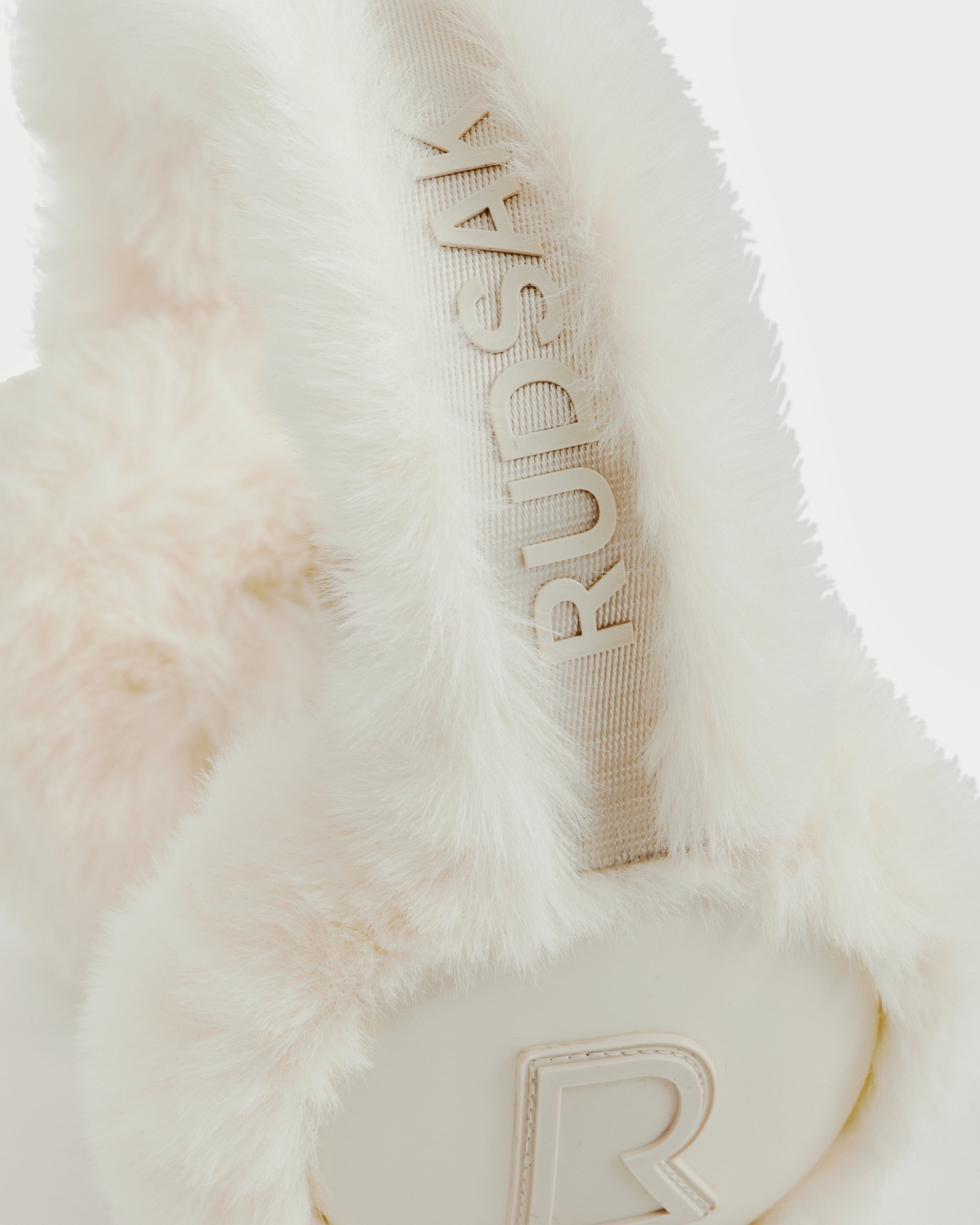 Close-up of VALENCIA - 8425543 Iceland white faux fur earmuffs by Rudsak, a chic winter accessory with the brand name embossed on the headband and a raised “R” logo on the ear cover.