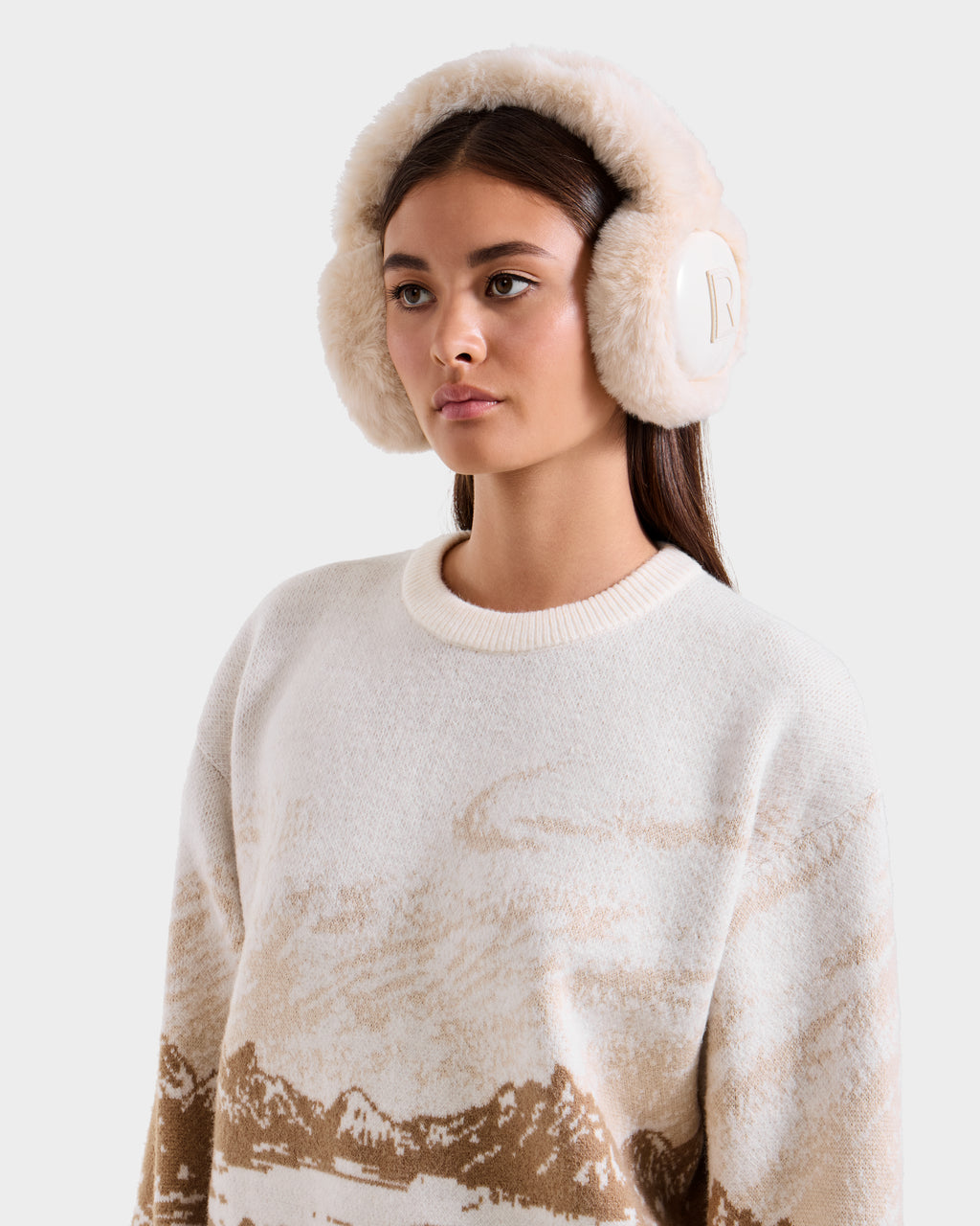 A woman with long brown hair wears Rudsak VALENCIA - 8425543 Vanilla fluffy faux fur earmuffs and a cream sweater with a tan mountain landscape pattern. She stands against a plain light background, looking ahead neutrally.