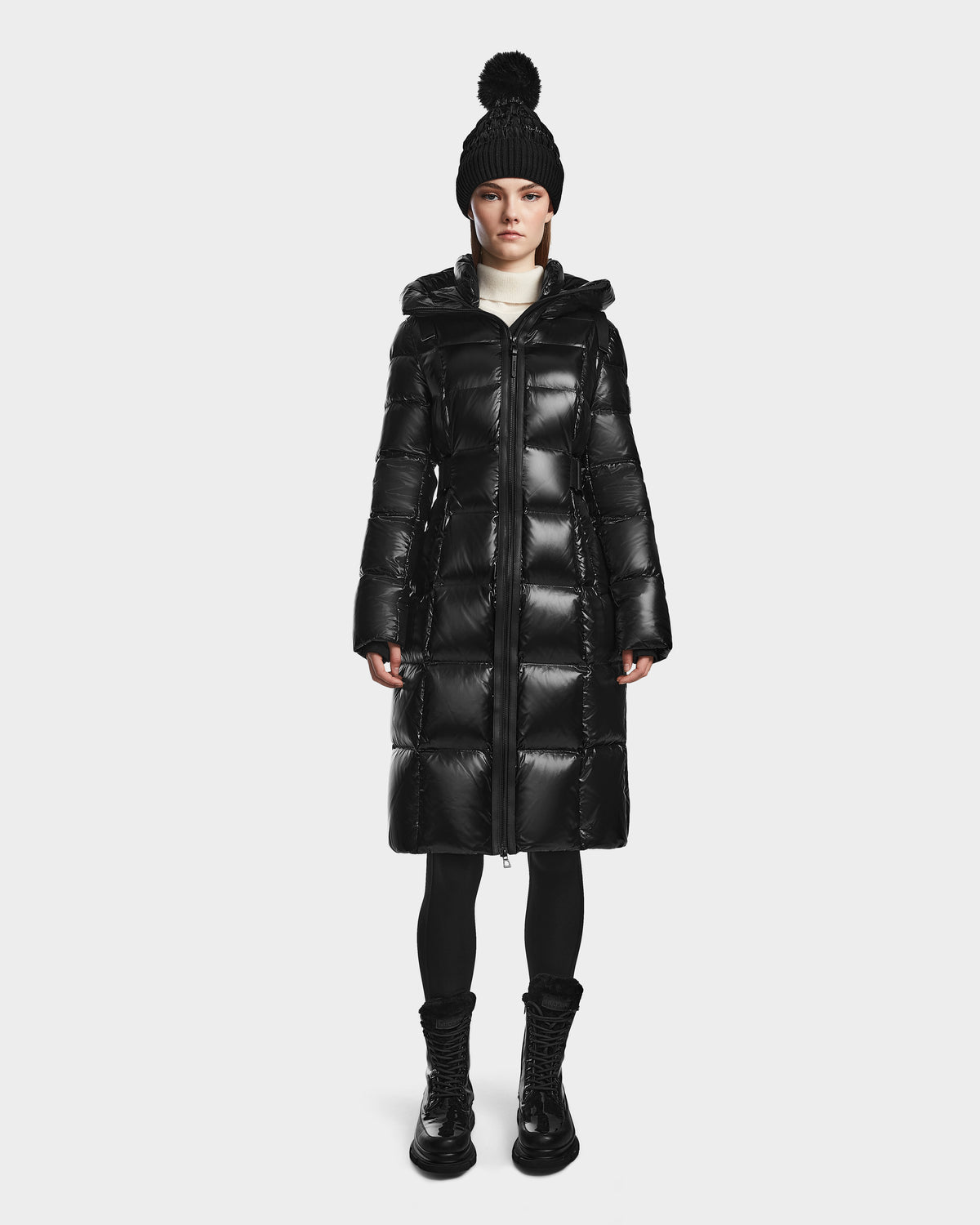 A woman faces forward on a plain white background, wearing Rudsak's VENICE S - L124514 Black long quilted down puffer coat (RDS-certified), paired with a black pom-pom beanie, leggings, and boots.