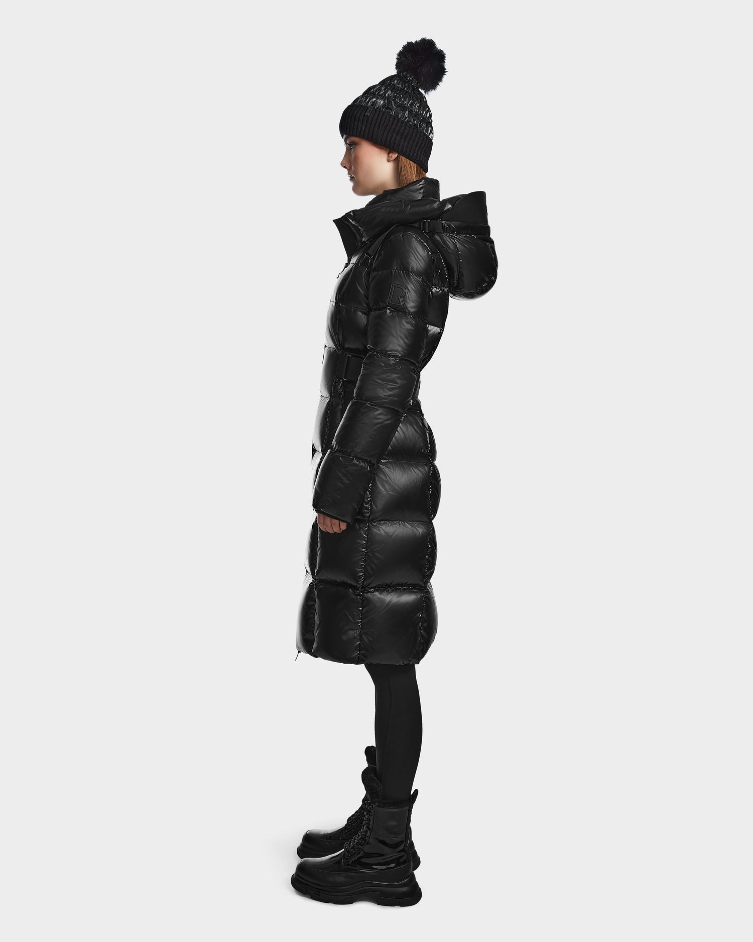 A person stands in profile against a plain white background, wearing the Rudsak VENICE S - L124514 Black women's winter coat with a hood, paired with black leggings, chunky black boots, and a black knit beanie with pom-pom.
