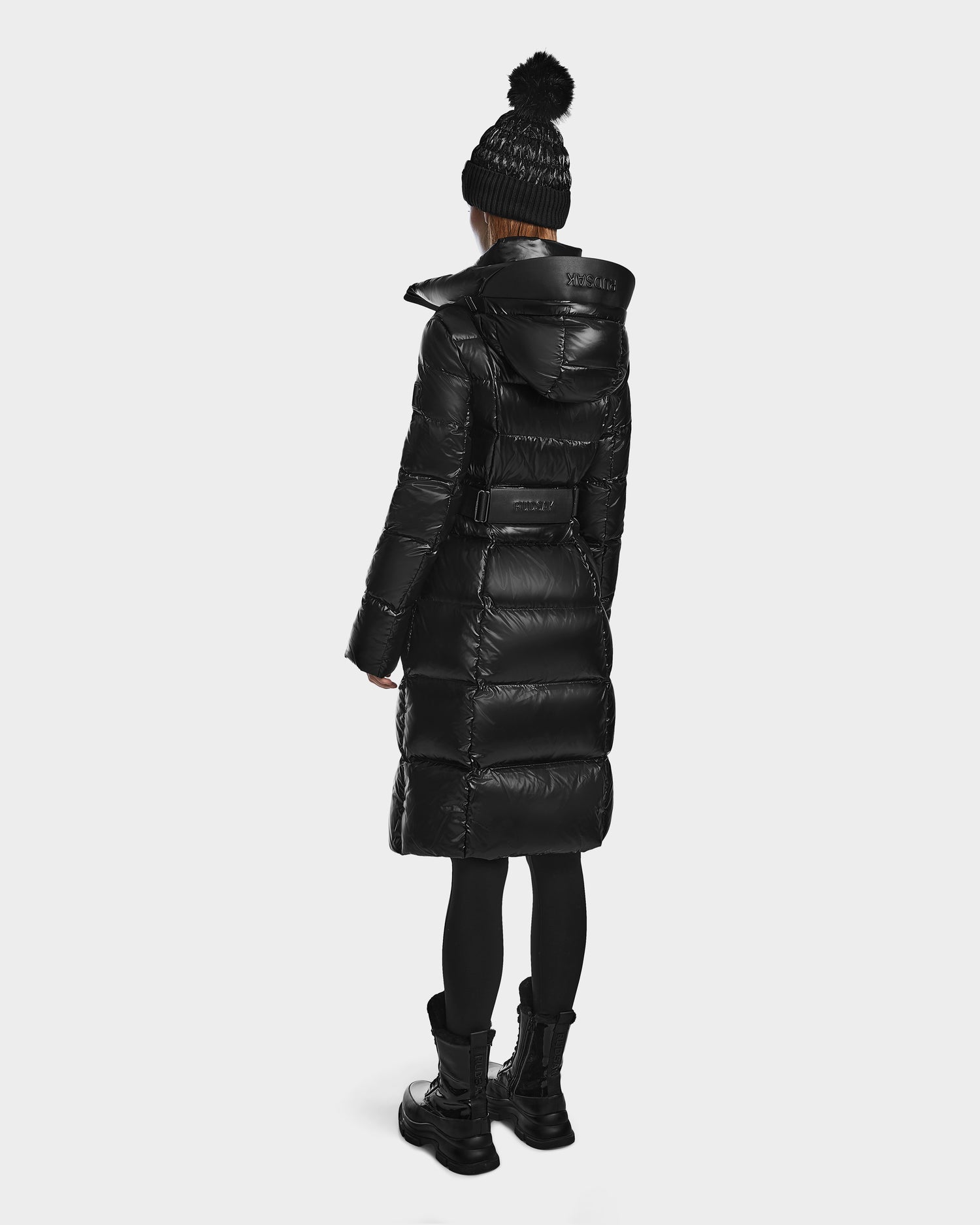 A person wears the VENICE S - L124514 Black puffer by Rudsak, paired with a black knit pom-pom hat, black leggings, and boots, standing facing away against a plain white background.