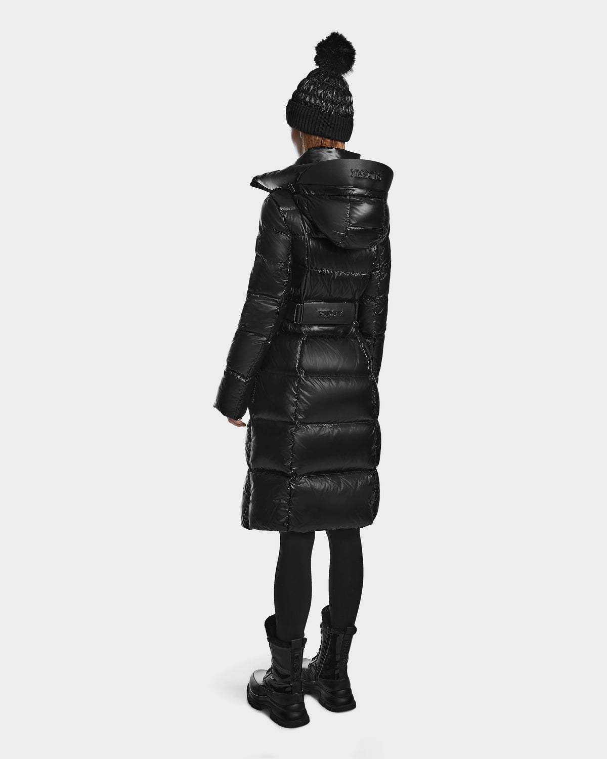 A person wears the VENICE S - L124514 Black puffer by Rudsak, paired with a black knit pom-pom hat, black leggings, and boots, standing facing away against a plain white background.