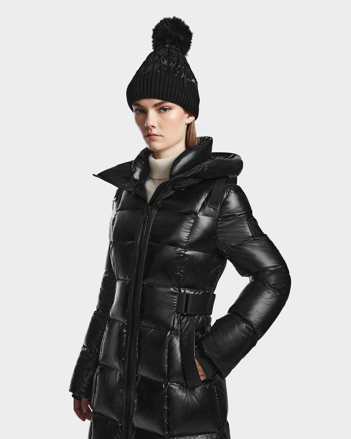 A woman wears the Rudsak VENICE S - L124514 Black long quilted down puffer coat and a black knit pom-pom beanie, standing with hands in her pockets and looking ahead against a plain background.