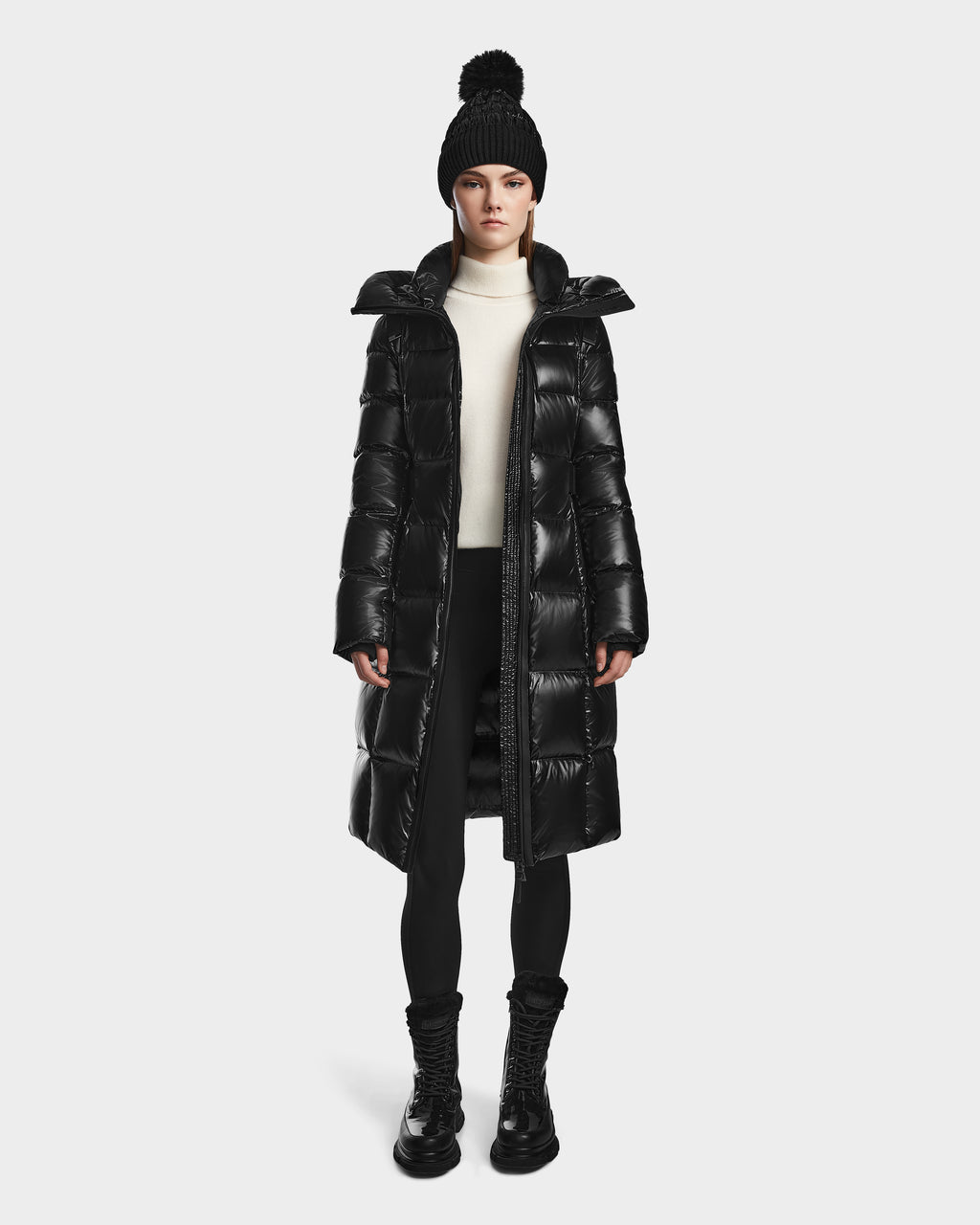 A woman wearing the Rudsak VENICE S - L124514 Black winter coat, a black pom-pom knit hat, white turtleneck, black leggings, and boots stands facing forward against a plain white background. The quilted puffer features RDS-certified down.