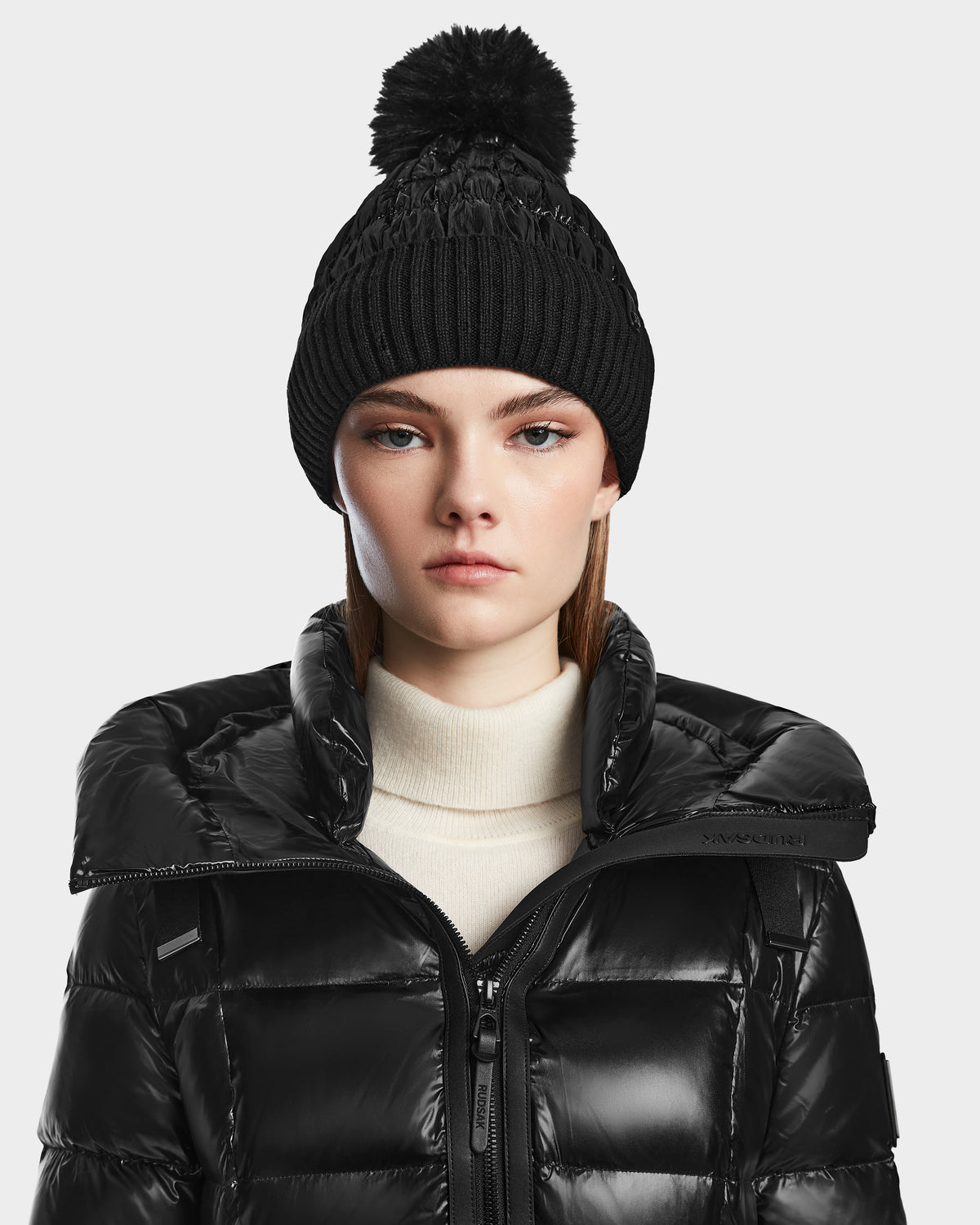A woman wears the Rudsak VENICE S - L124514 Black down puffer jacket with a black knit beanie featuring a pom-pom and a light turtleneck, looking directly at the camera with a neutral expression.