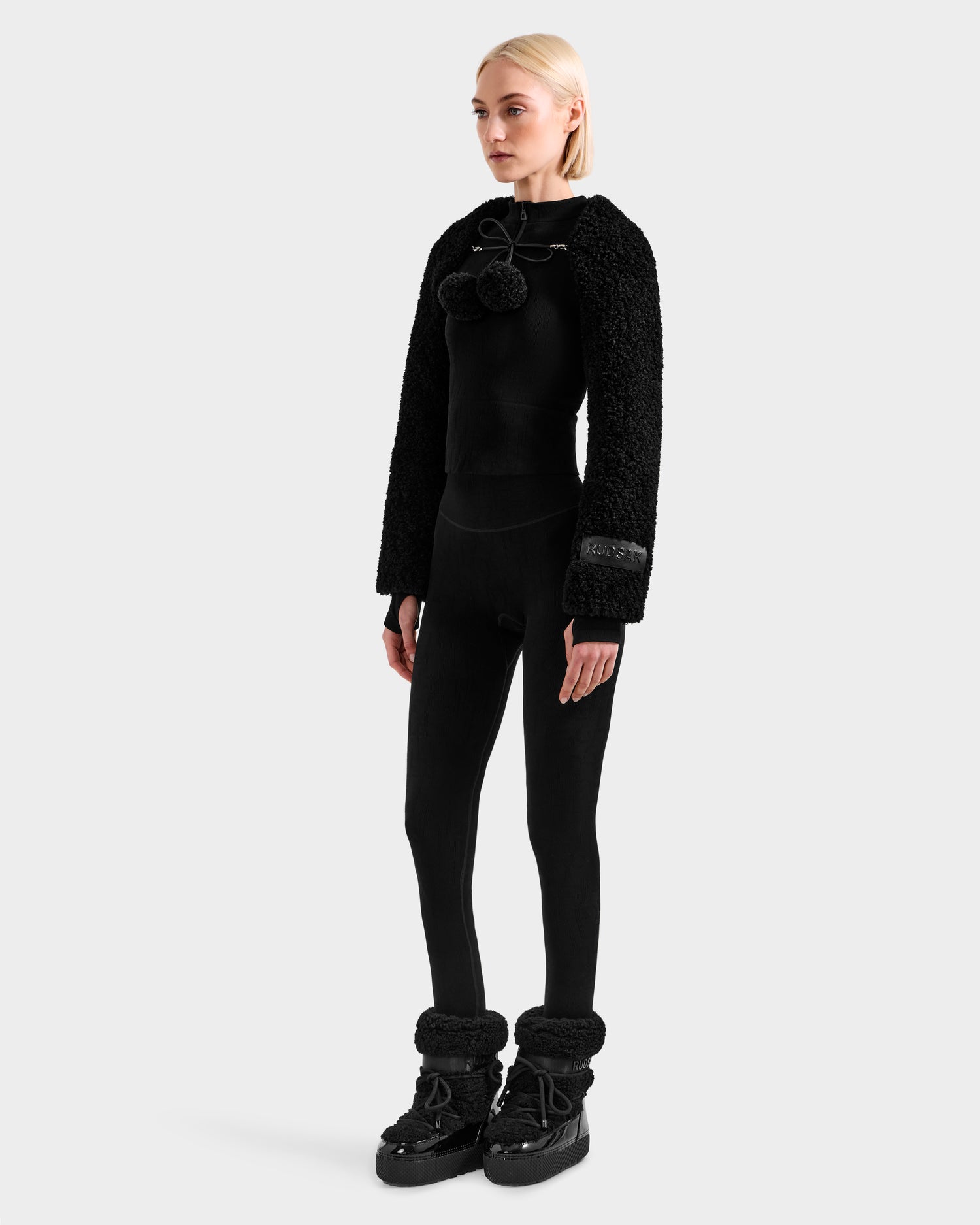 A blonde woman wears the Rudsak VERITY - 8525666 Black textured jacket with a black fitted top, leggings, and fur-trimmed boots, posing against a plain light gray background.