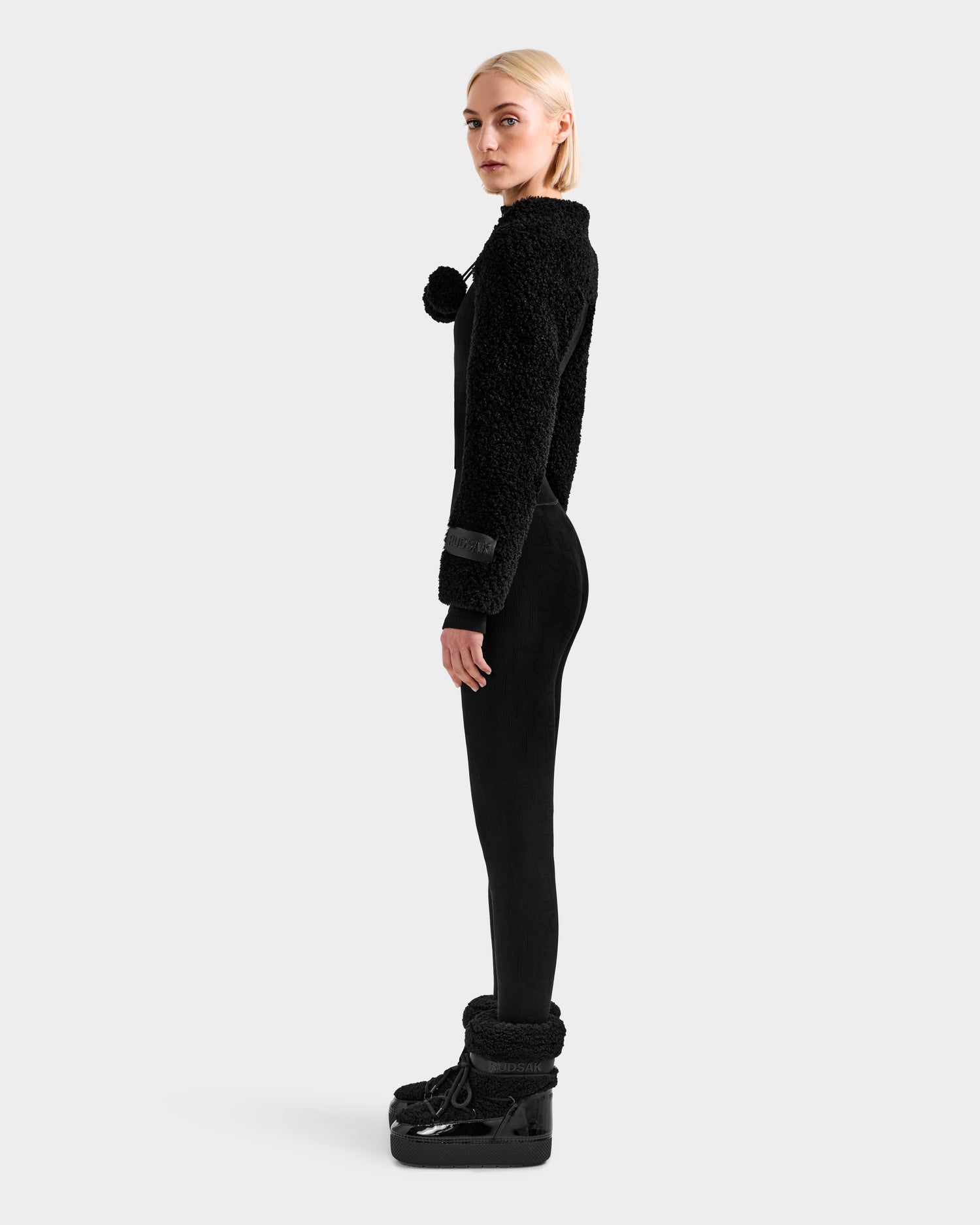 A woman with blonde hair stands in profile wearing Rudsak's VERITY - 8525666 Black fuzzy sweater with a pom-pom, paired with black leggings and winter boots, set against a plain light gray background.