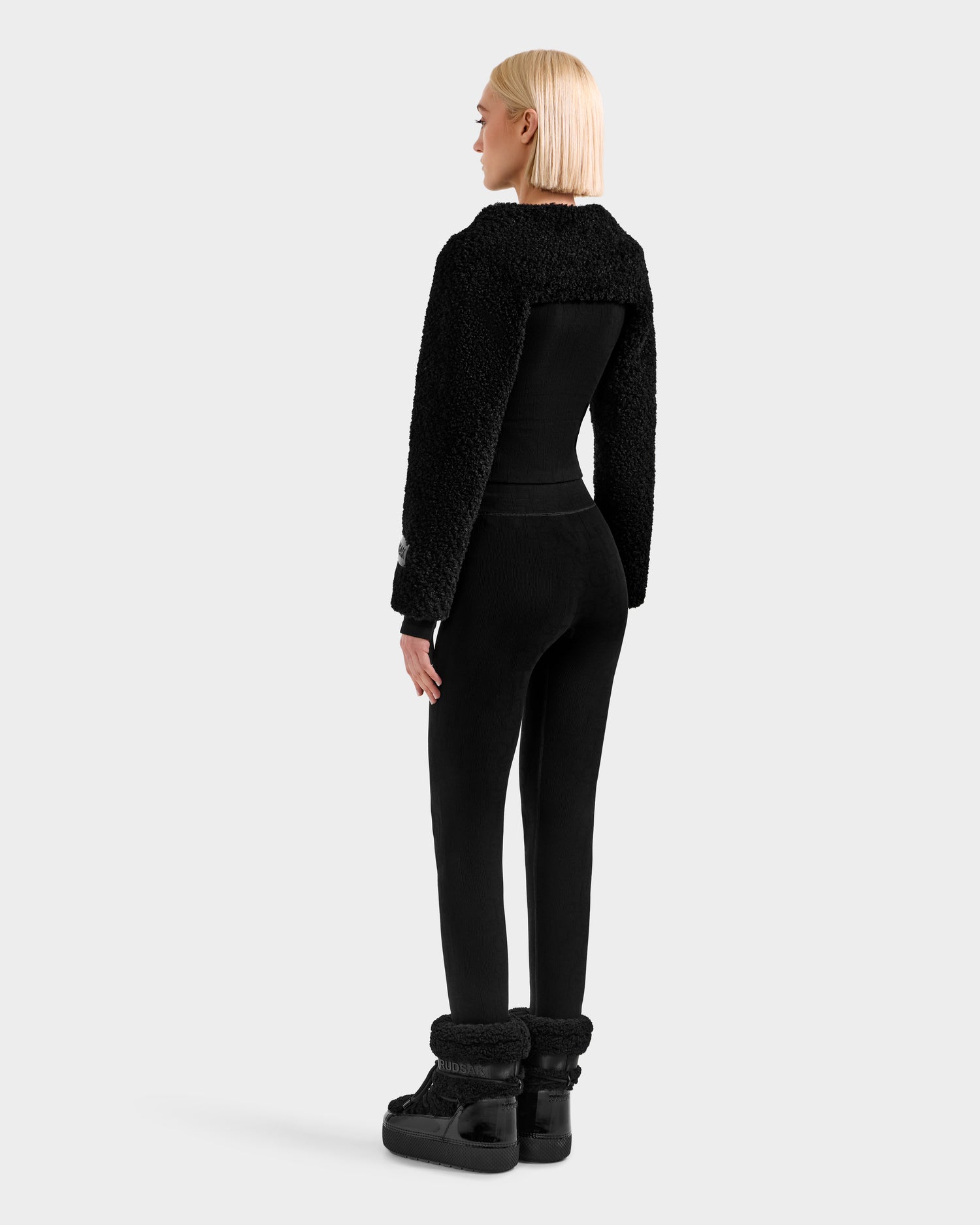 A woman with blonde hair faces away, wearing the Rudsak VERITY - 8525666 Black fitted outfit with textured sleeves, black pants, platform boots, and a chic women’s faux shearling bolero—set against a light gray background.