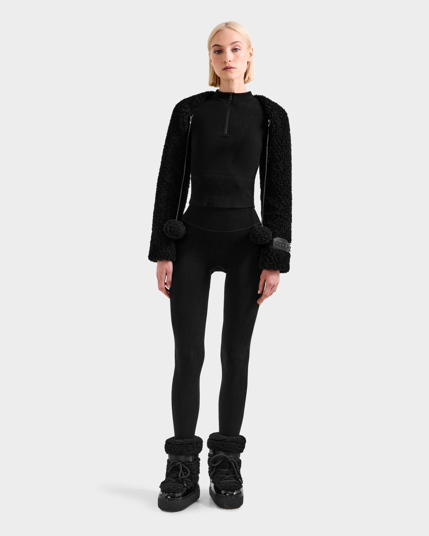 A woman faces forward in the Rudsak VERITY - 8525666 Black women’s faux shearling bolero jacket with pom-pom details, styled with a black fitted zip-up top, black leggings, and winter boots against a light gray background.