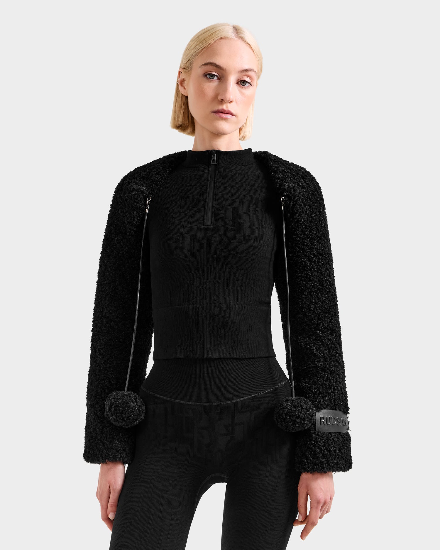 A woman with straight blonde hair wears the Rudsak VERITY - 8525666 Black top, featuring long textured sleeves and pom-pom zipper details, standing against a light gray background and looking directly at the camera.