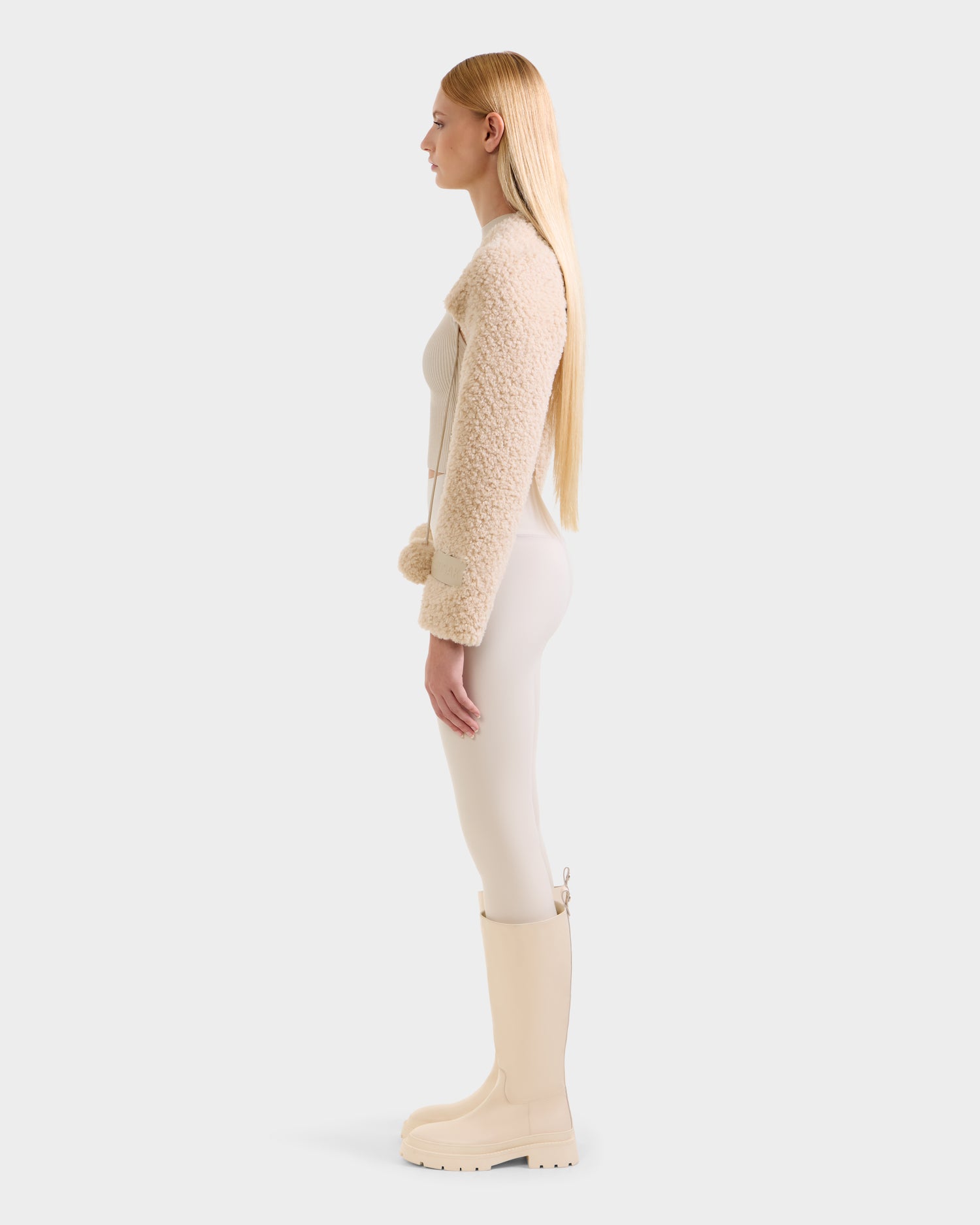 A woman with long blonde hair stands in profile wearing the Rudsak VERITY - 8525666 Vanilla cream-textured jacket, light pants, and matching knee-high boots against a plain, light background.