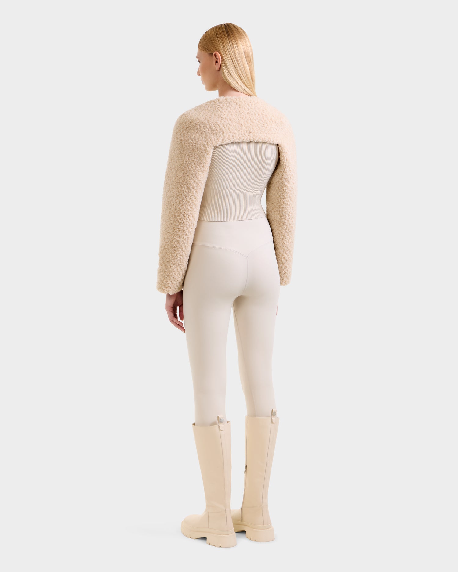 A person stands facing away in the Rudsak VERITY - 8525666 Vanilla cropped, textured beige faux shearling bolero, styled with fitted light beige leggings, beige knee-high boots, and straight blonde hair. The background is light gray.