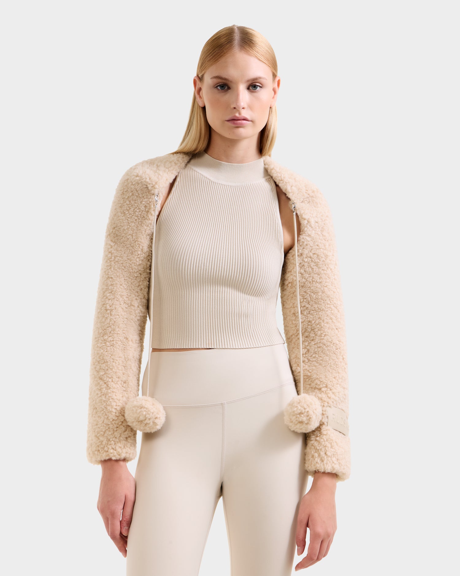 A woman with straight blonde hair wears the Rudsak VERITY - 8525666 Vanilla faux shearling bolero with pom-pom ties over a beige sleeveless top and high-waisted pants, standing against a plain light background.