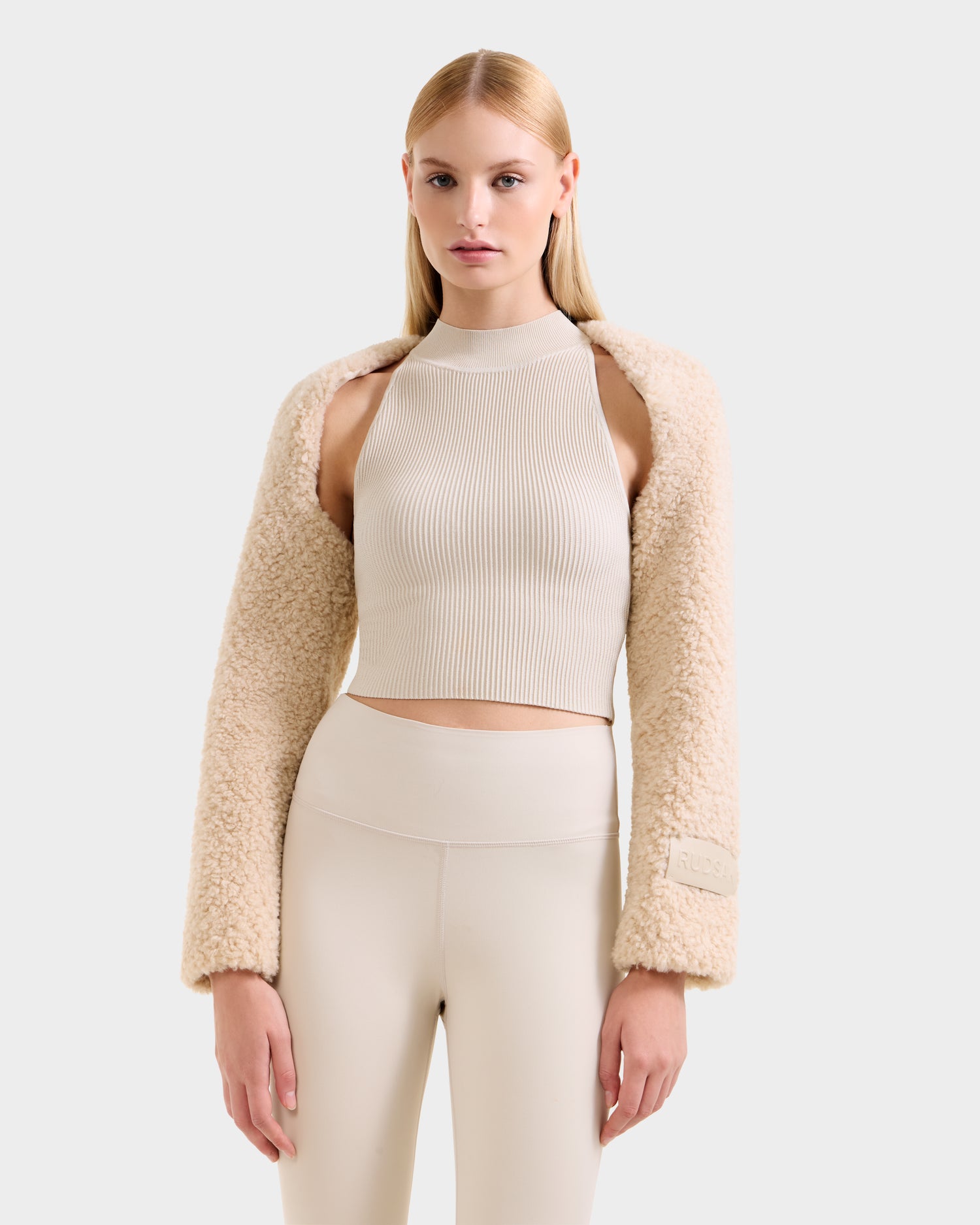 A woman with straight blonde hair wears the Rudsak VERITY - 8525666 Vanilla faux shearling bolero over a beige ribbed crop top and high-waisted fitted pants, posing against a plain light gray background with a neutral expression.