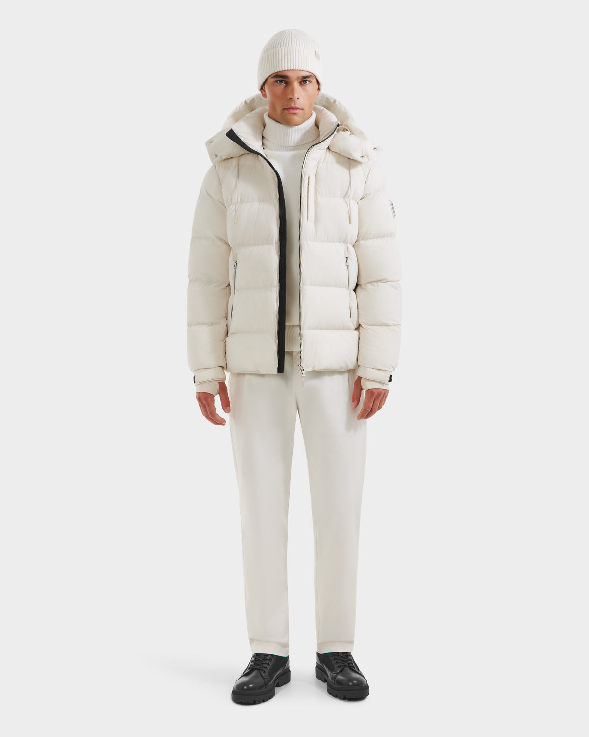 A man stands facing forward, wearing the Rudsak VINCE M - 6124561 Vanilla men’s down puffer jacket, a white turtleneck, pants, beanie, and black shoes against a plain light background.