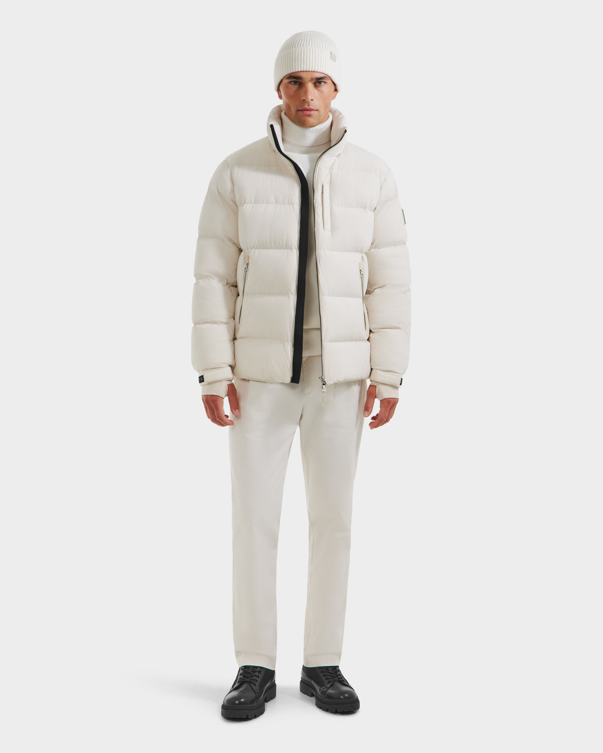 A man faces forward in an all-white outfit, featuring the Rudsak VINCE M - 6124561 Vanilla puffer jacket, a beanie, turtleneck, and pants. He pairs this look with black shoes against a plain white background.