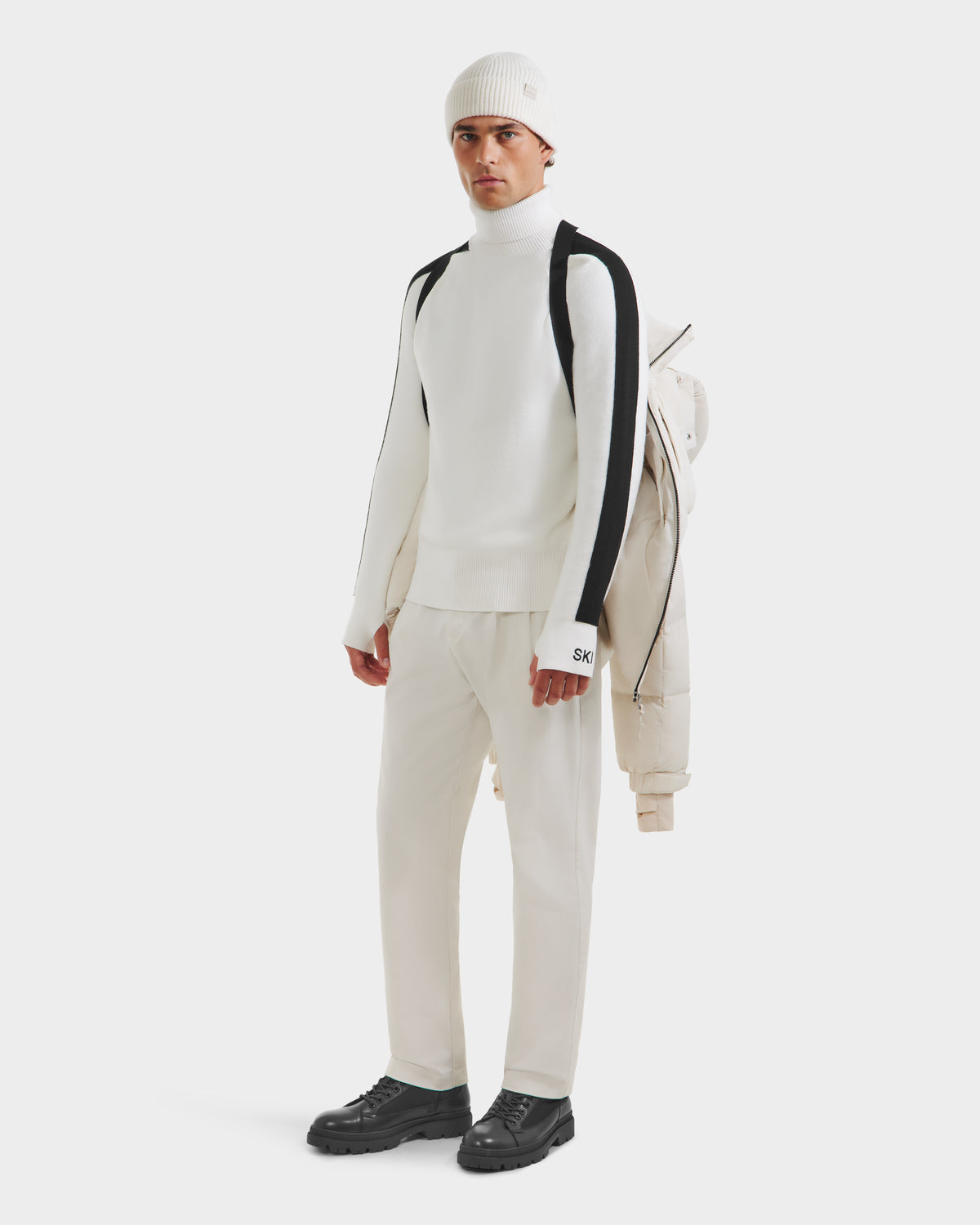 A man stands against a plain white background, holding the Rudsak VINCE M - 6124561 Vanilla weather-resistant jacket over his shoulder. He wears a white turtleneck with black stripes, white pants, a white beanie, and black shoes.