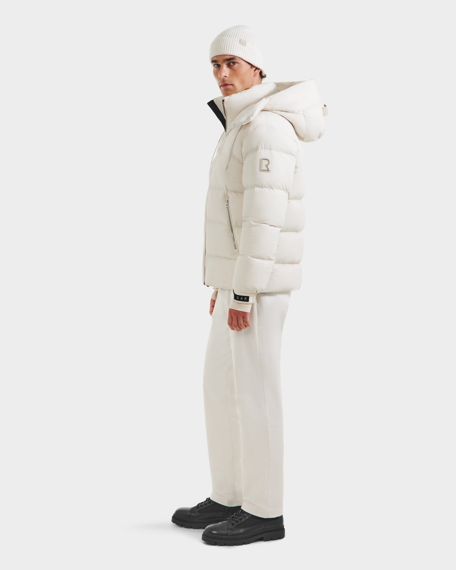A person wears the Rudsak VINCE M - 6124561 Vanilla mens down puffer jacket with matching pants and a beanie, standing slightly to the side in black shoes against a plain white background.