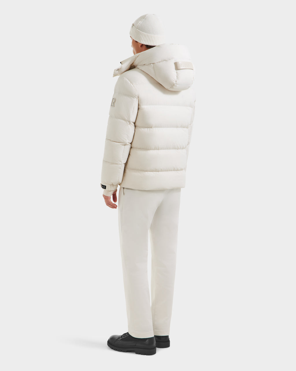 A person stands facing away, wearing the Rudsak VINCE M - 6124561 Vanilla men’s down puffer jacket with a hood, matching pants, a white knit beanie, and black shoes against a plain white background.