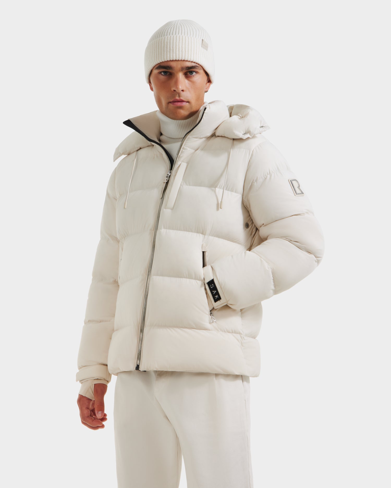 A man wears the Rudsak VINCE M - 6124561 Vanilla, a men's quilted down puffer jacket with RDS-certified down, plus a white beanie, turtleneck, and pants, standing facing forward against a plain white background.
