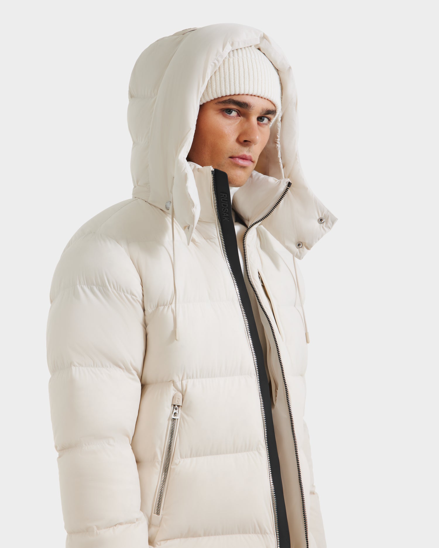 A person in a Rudsak VINCE M - 6124561 Vanilla mens down puffer jacket with hood and white knit beanie stands against a plain white background, looking slightly to the side. The weather-resistant jacket features RDS-certified down.