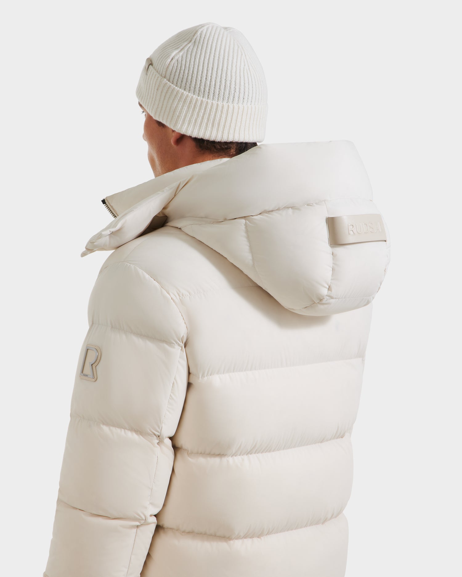 A person wearing the Rudsak VINCE M - 6124561 Vanilla men’s down puffer jacket with a hood and matching knit beanie, viewed from the back and slightly to the side against a plain white background.