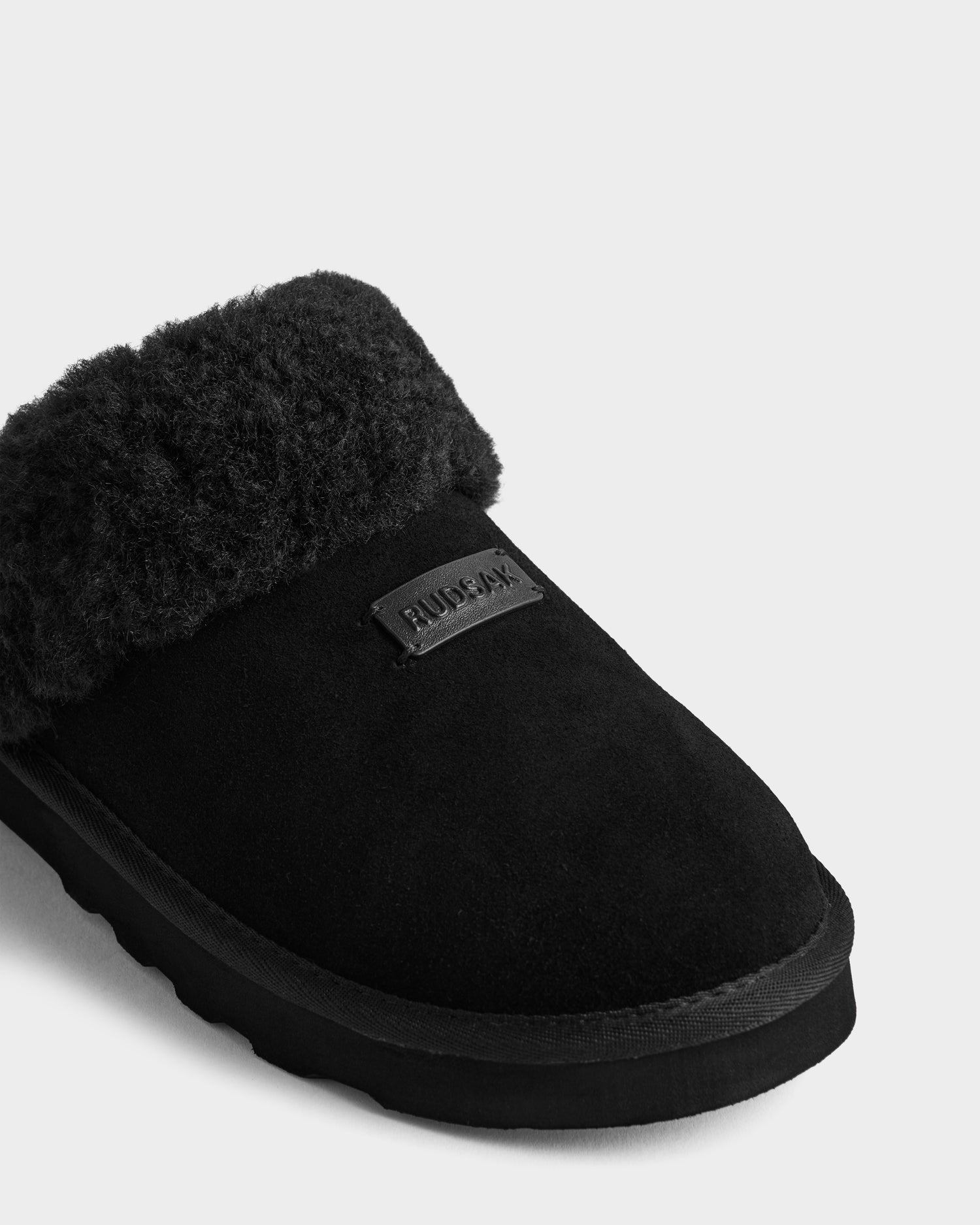 A close-up of the Rudsak WILBER - 6225509 Black suede slipper features a fluffy faux shearling cuff and a branded label on the upper, set against a plain light gray background.