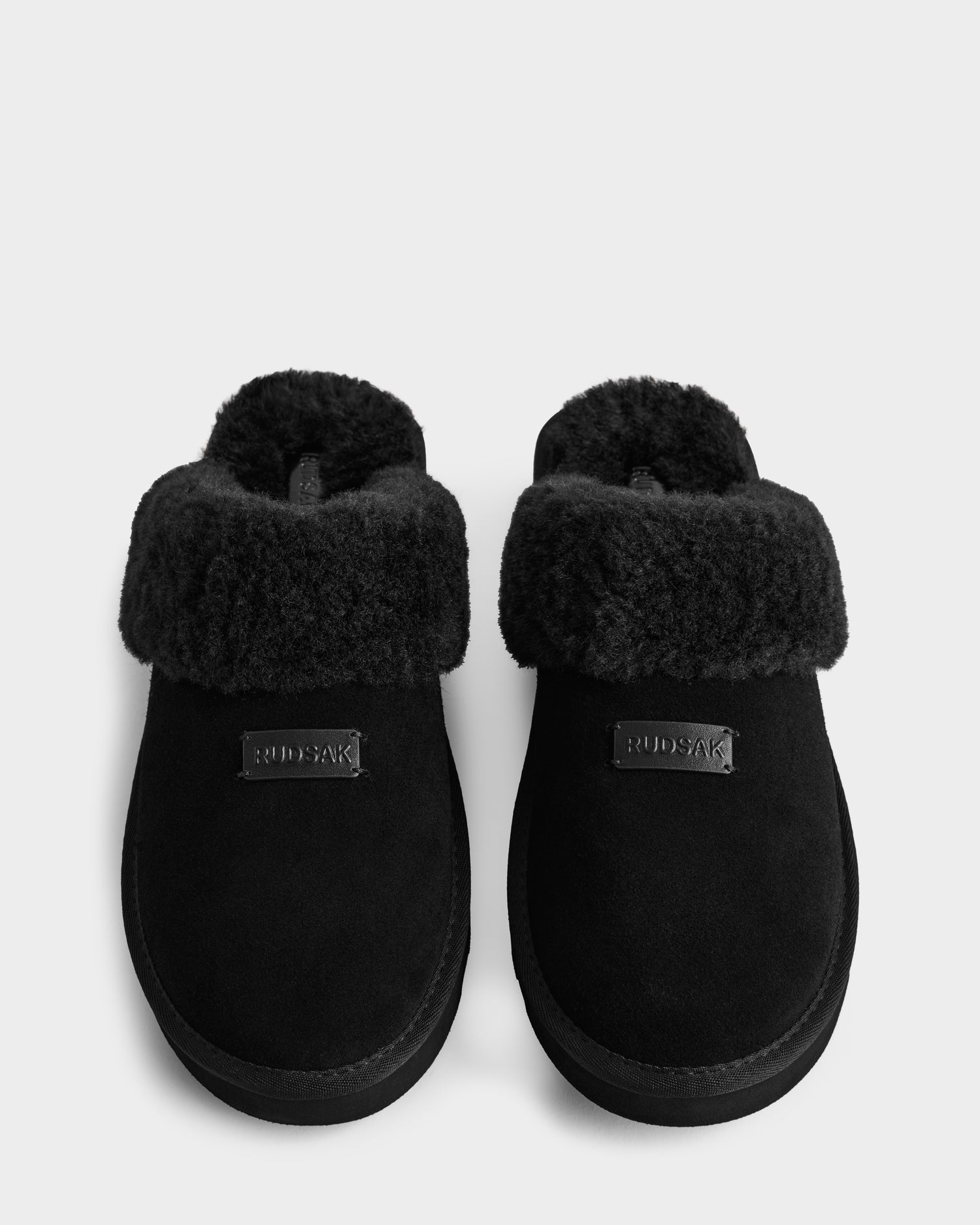 The WILBER - 6225509 Black slippers by Rudsak feature plush faux shearling cuffs, a soft suede texture, and a small front label. This stylish men's pair is shown side by side on a white background.