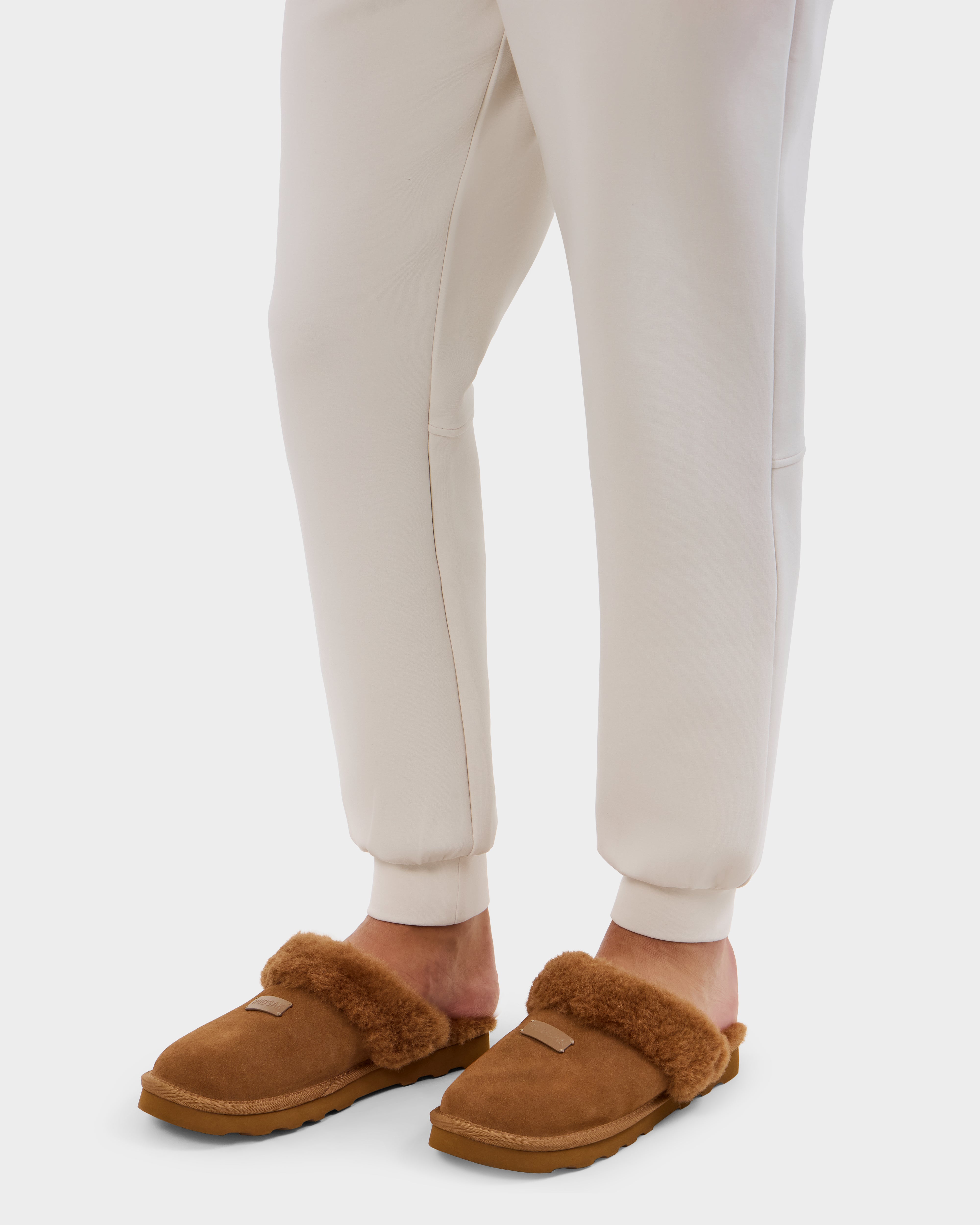 A person wearing Rudsak's WILBER - 6225509 Tan faux shearling slippers and cream jogger pants stands on a white background.