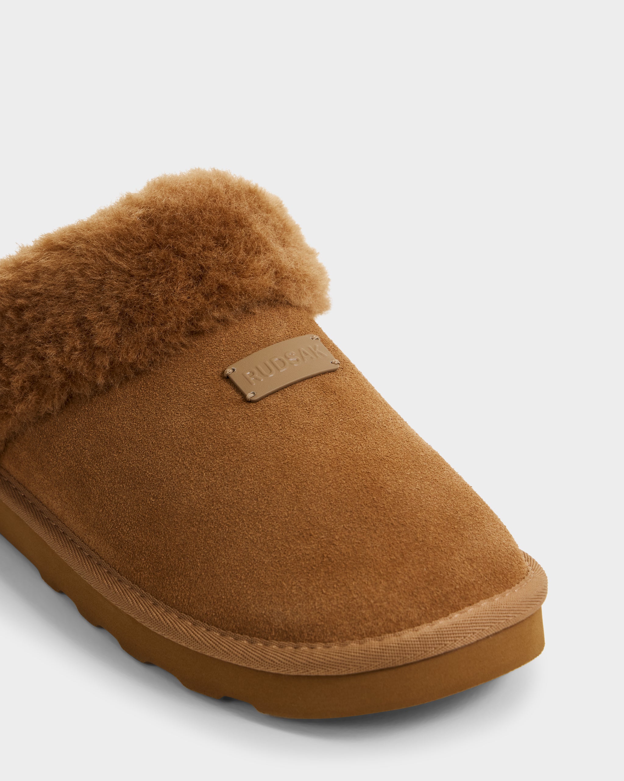 A close-up of the WILBER - 6225509 Tan men's suede slipper by Rudsak, featuring a fluffy textured collar, rubber sole, and a small rectangular label on the upper marked RUDENY.