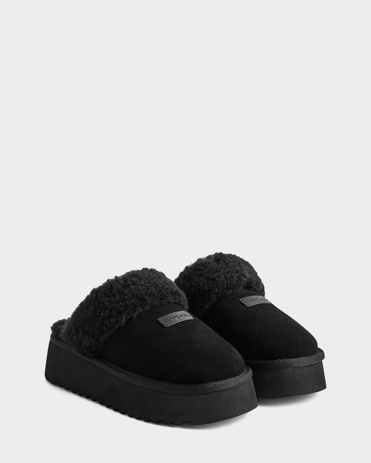 The Rudsak WILMA - 8225582 Black women's slipper slip-ons feature plush fuzzy trim, thick soles, and a patch label on the front—perfect for cozy winter style. Shown against a plain light gray background.
