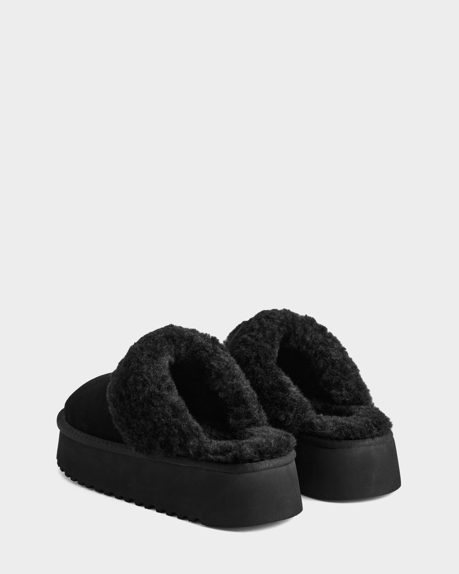 The Rudsak WILMA - 8225582 Black women’s suede slippers feature thick, textured soles and plush faux shearling linings. Displayed on a light gray background, the slippers are positioned slightly facing away from each other.