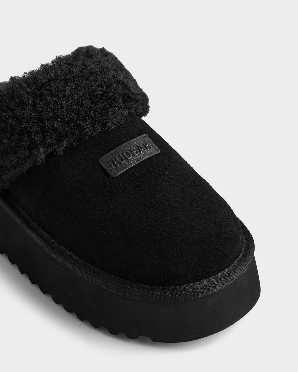 Close-up of the WILMA - 8225582 Black women’s slipper by Rudsak, made from soft suede with fuzzy faux shearling lining, thick sole, and a stitched rectangular RUDSAK patch on top. The background is plain and light-colored.