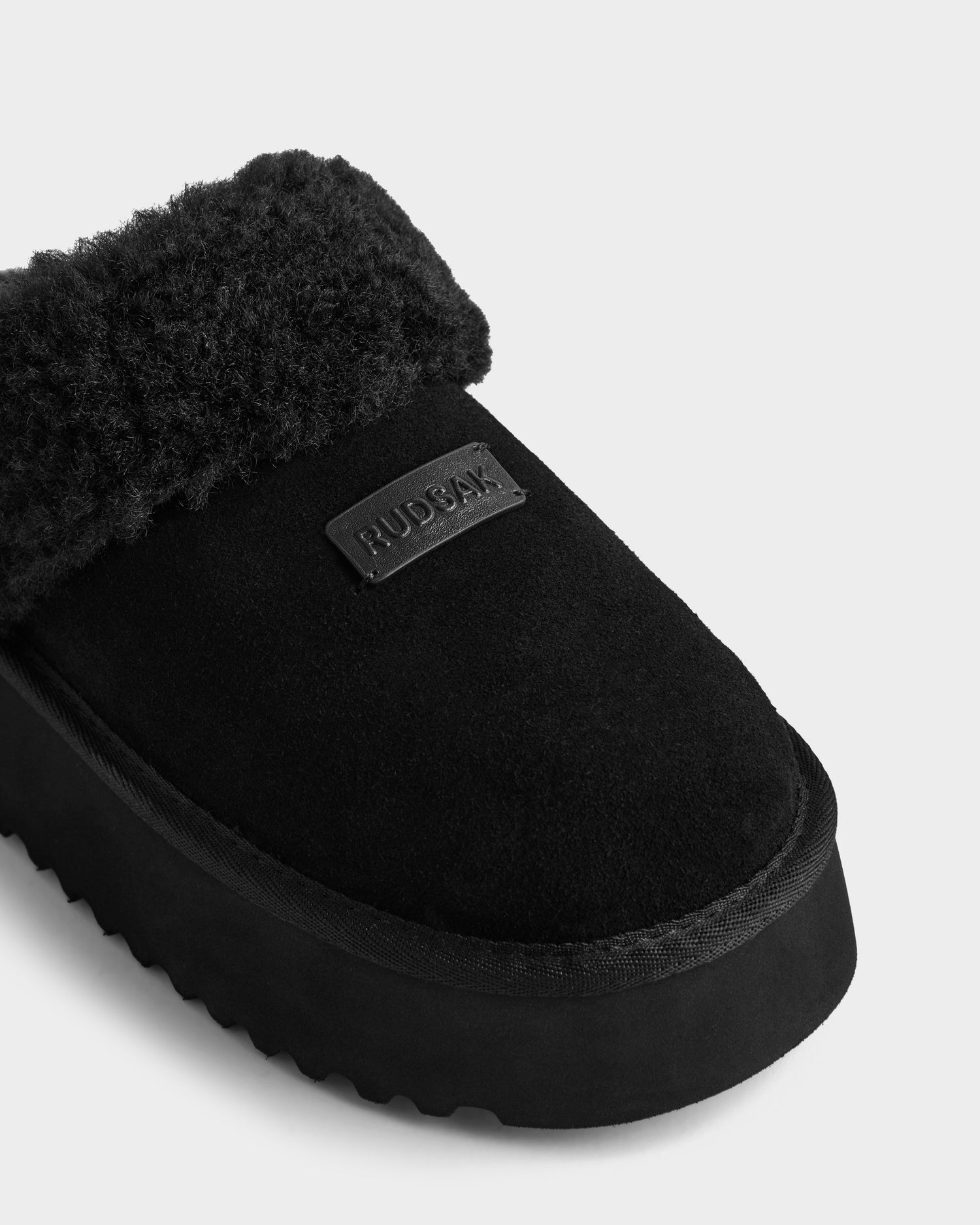 Close-up of the WILMA - 8225582 Black women’s slipper by Rudsak, made from soft suede with fuzzy faux shearling lining, thick sole, and a stitched rectangular RUDSAK patch on top. The background is plain and light-colored.