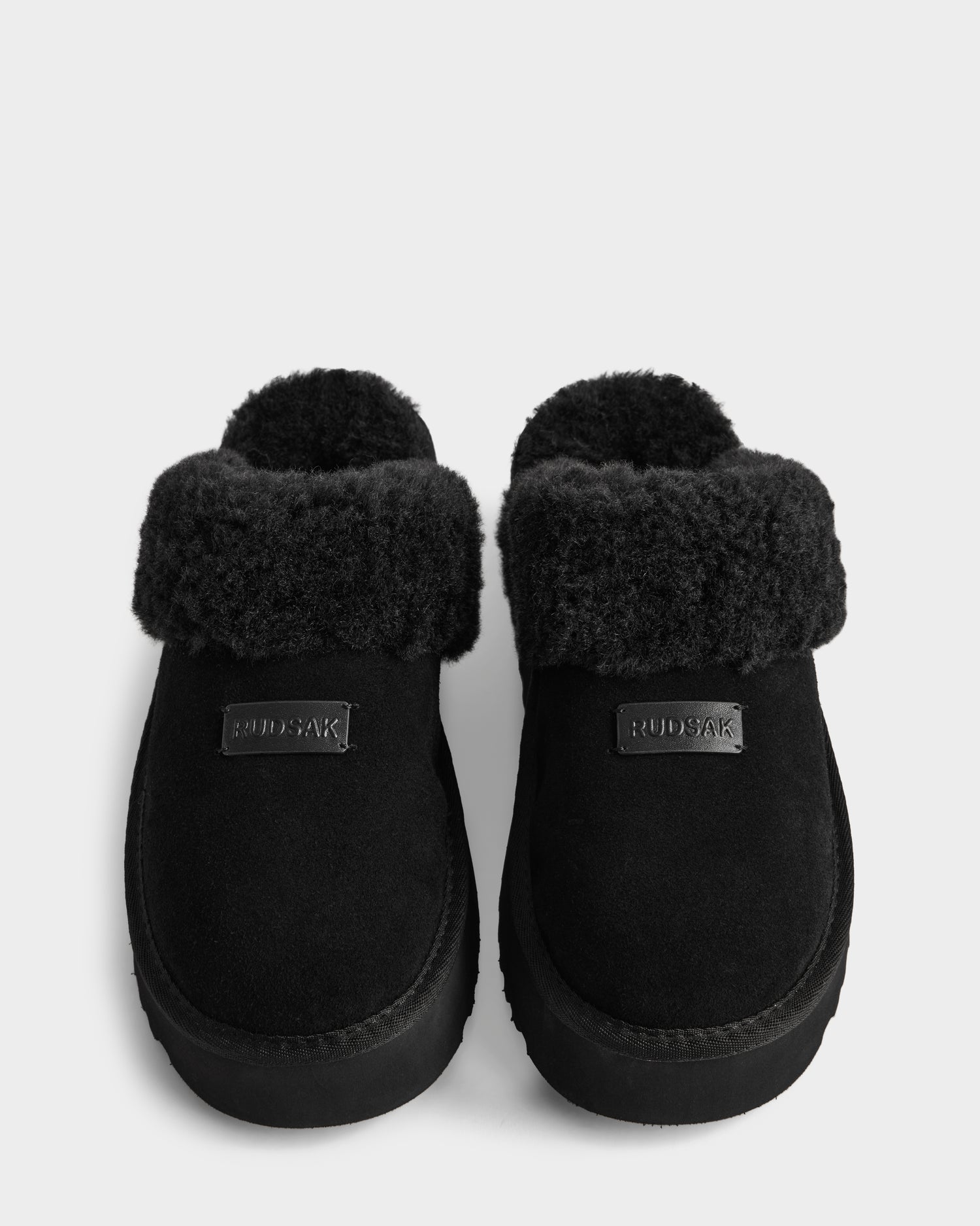 The WILMA - 8225582 Black women’s slippers by Rudsak feature fluffy, folded faux shearling cuffs, a suede-like texture, and the Rudsak logo on each slipper, shown on a plain white background.