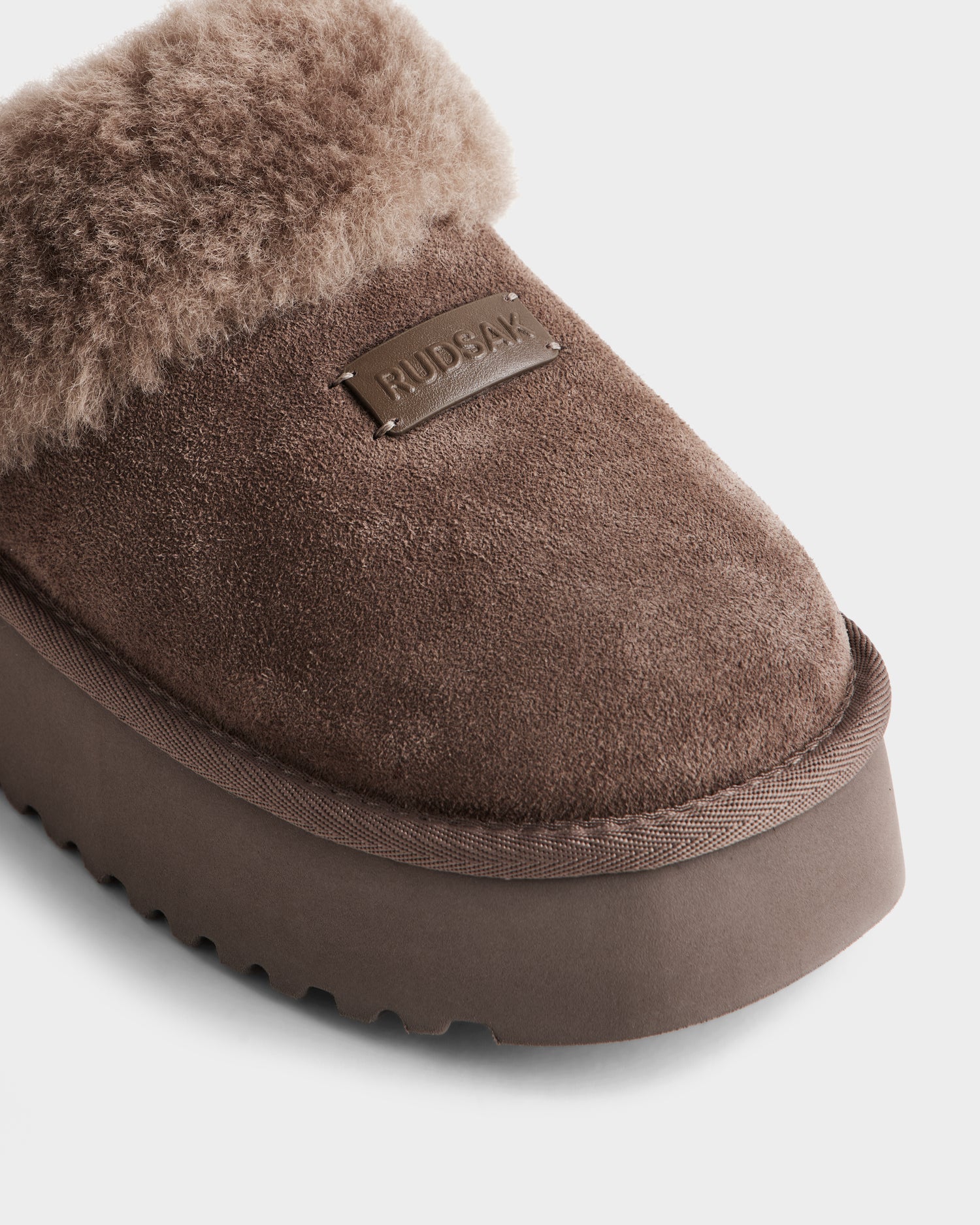Close-up of Rudsak's WILMA - 8225582 Mocha women’s suede slipper, featuring a fuzzy faux shearling cuff, thick rubber sole, and a small rectangular metal plate engraved with RUDSAK on the upper.