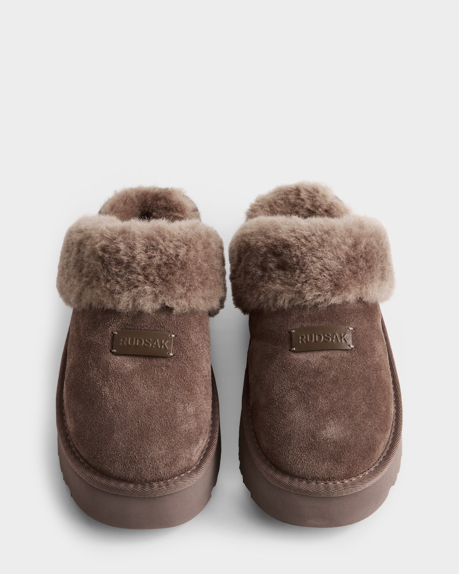 A pair of Rudsak WILMA - 8225582 Mocha women’s brown suede slippers with plush faux shearling lining, folded-over cuff, and RUDSAK labels on the front uppers, shown against a white background.