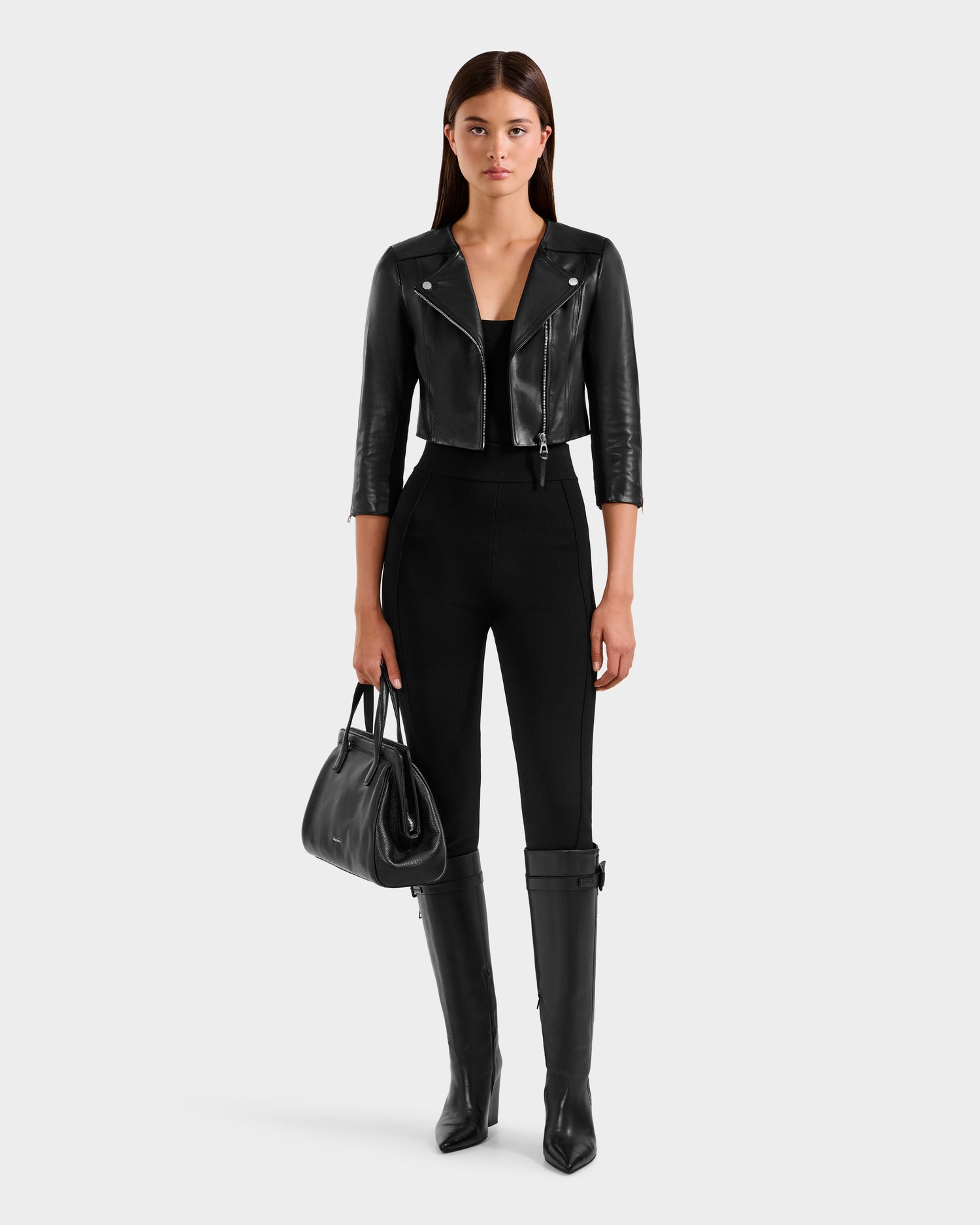 A woman with straight dark hair and a neutral expression wears the WINNI - 8124053 Black lamb leather biker jacket by Rudsak, styled with black pants, knee-high boots, and a handbag against a plain white background.