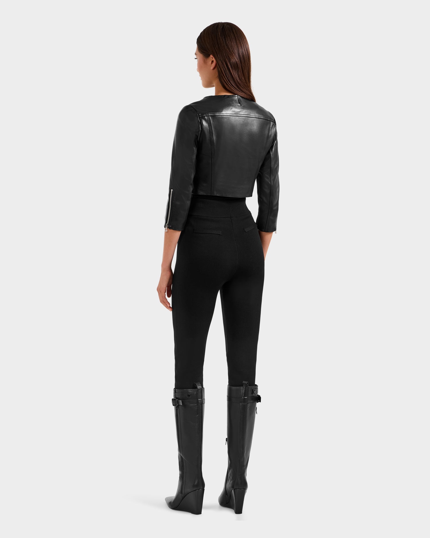 A woman faces away in the Rudsak WINNI - 8124053 Black cropped leather biker jacket, styled with high-waisted black pants and knee-high black buckle boots. Her straight brown hair is down against a plain light gray background.