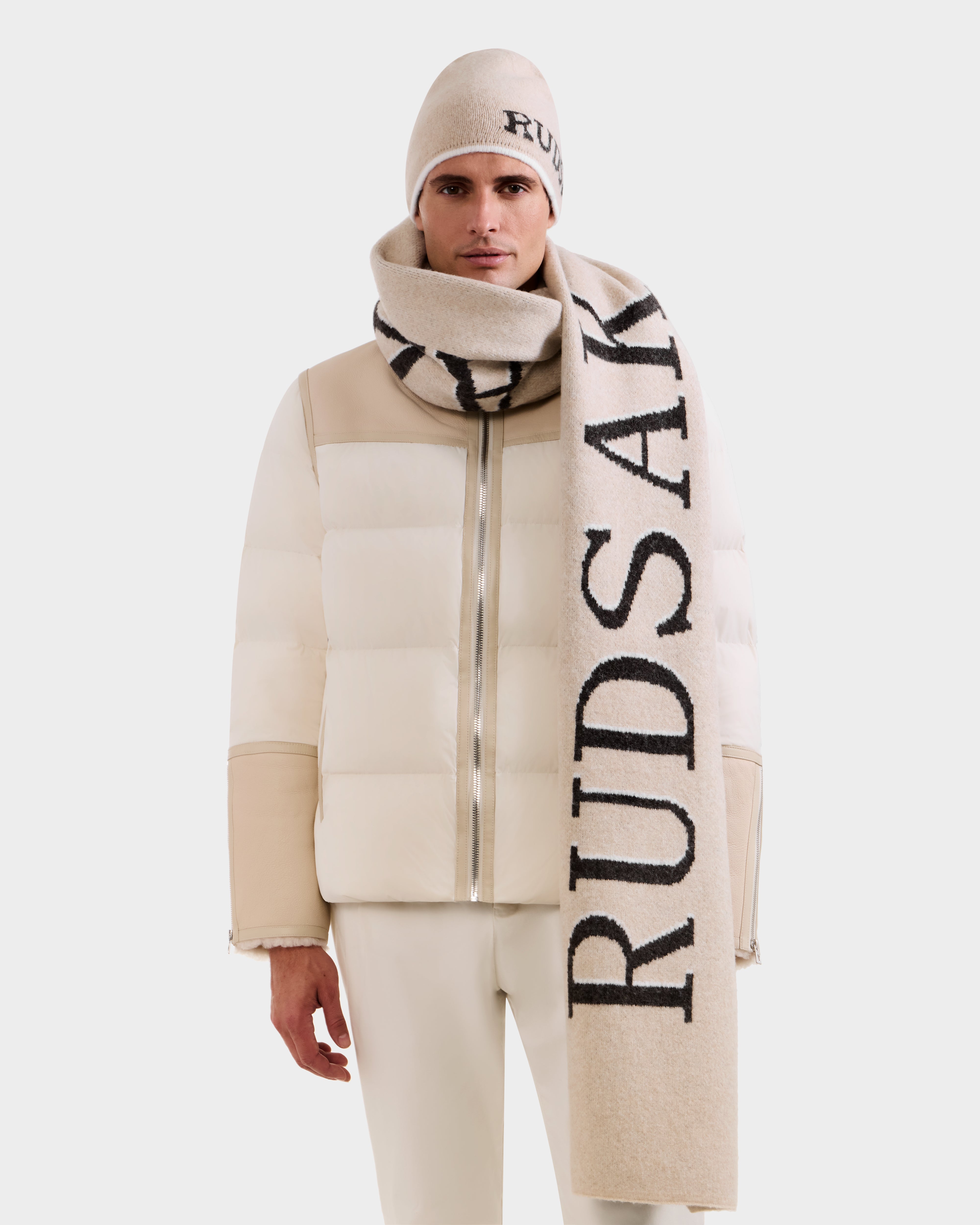 A man models Rudsak’s WYATT - 6425039 Vanilla puffer jacket with matching pants and a beige men’s scarf and beanie emblazoned with RUDSAK in bold black, standing against a plain white background.