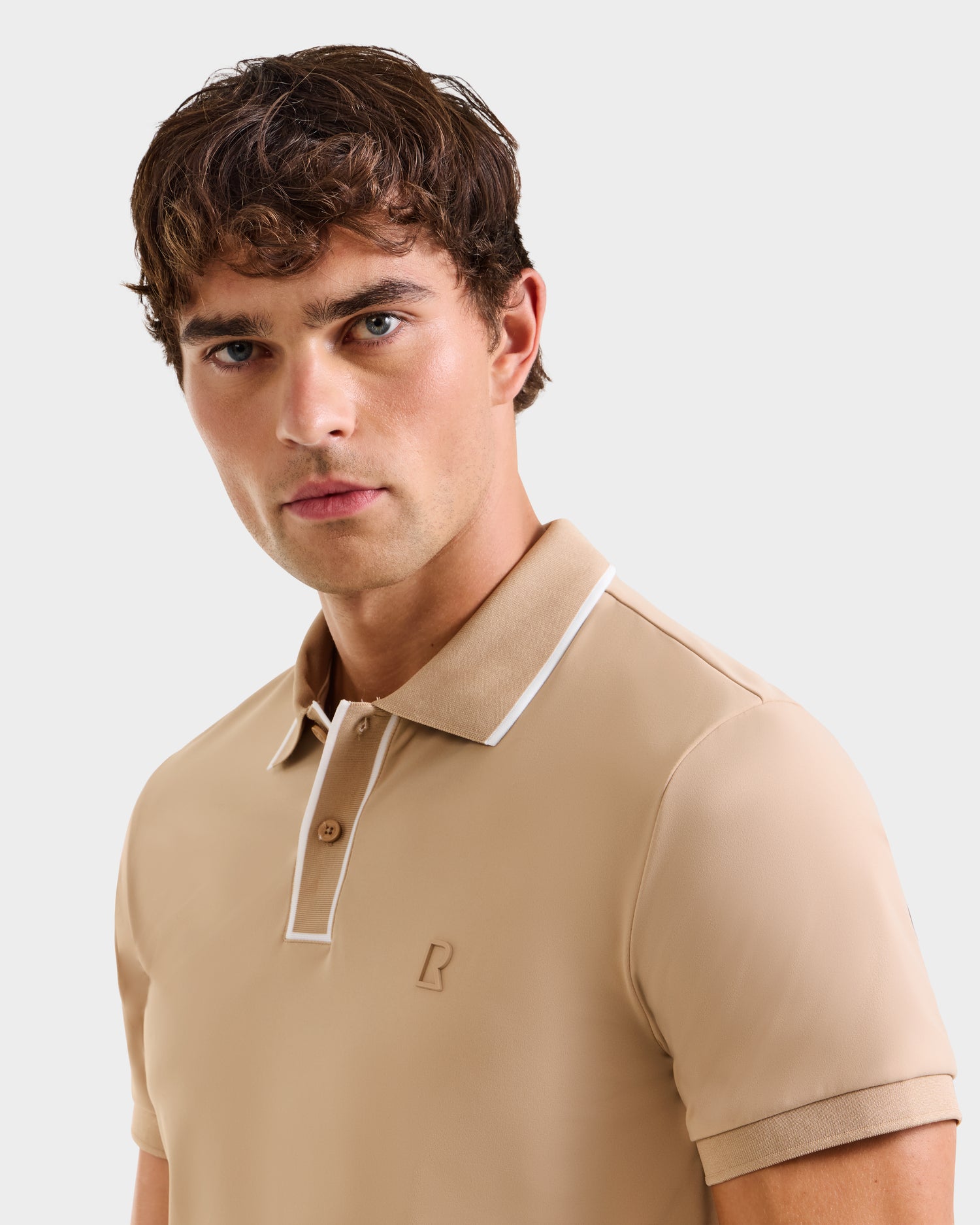 A young man with wavy brown hair wears the Rudsak XAVIER - 6524027 Almond men’s polo, beige with white trim on the collar and placket, and a small chest logo, set against a plain light background.
