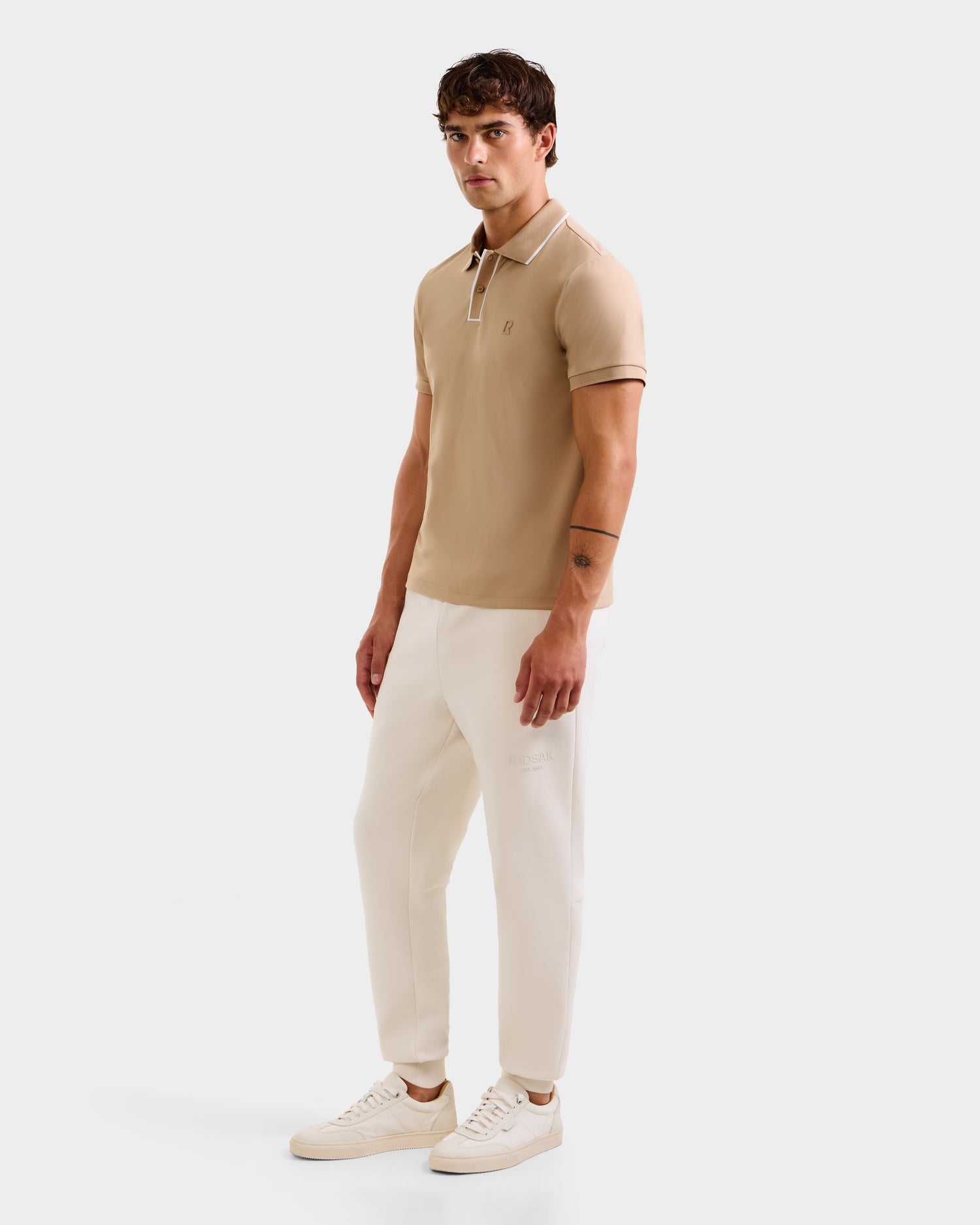A man with short brown hair and a neutral expression wears the XAVIER - 6524027 Almond polo shirt by Rudsak, styled with white pants and sneakers, standing against a plain white background with one arm by his side.