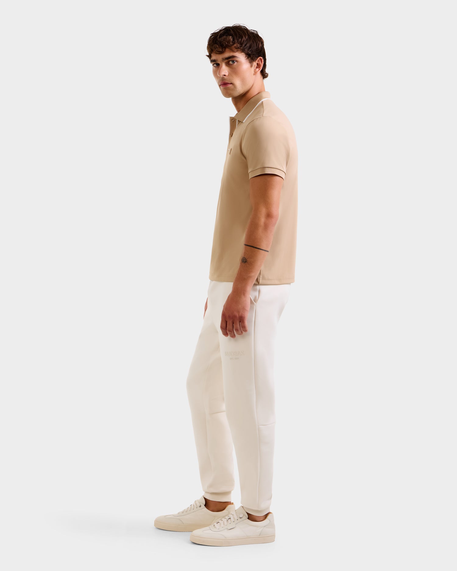 A man stands sideways against a plain white background wearing the Rudsak XAVIER - 6524027 Almond polo shirt, paired with white pants and white sneakers.