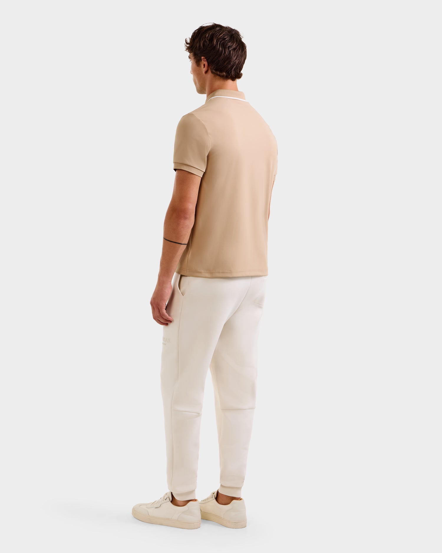 A man faces away, wearing the Rudsak XAVIER - 6524027 Almond mens polo shirt with white pants and matching almond sneakers, set against a plain white background.
