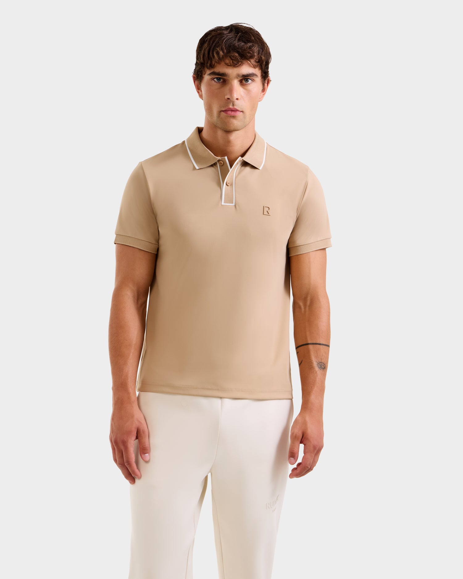 A man faces forward against a plain white background, wearing the Rudsak XAVIER - 6524027 Almond men's polo shirt in beige with white collar trim and off-white pants.