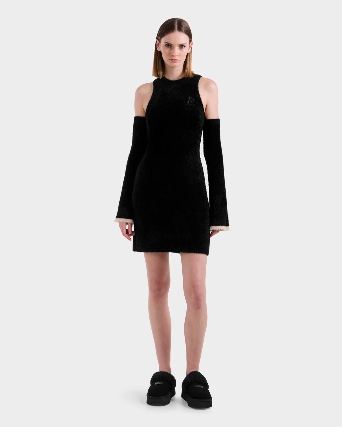 A woman stands facing forward in the Rudsak ZARAH - 8525696 Black fitted dress with cold-shoulder cutouts and detached long sleeves, paired with black platform shoes, against a plain white background.