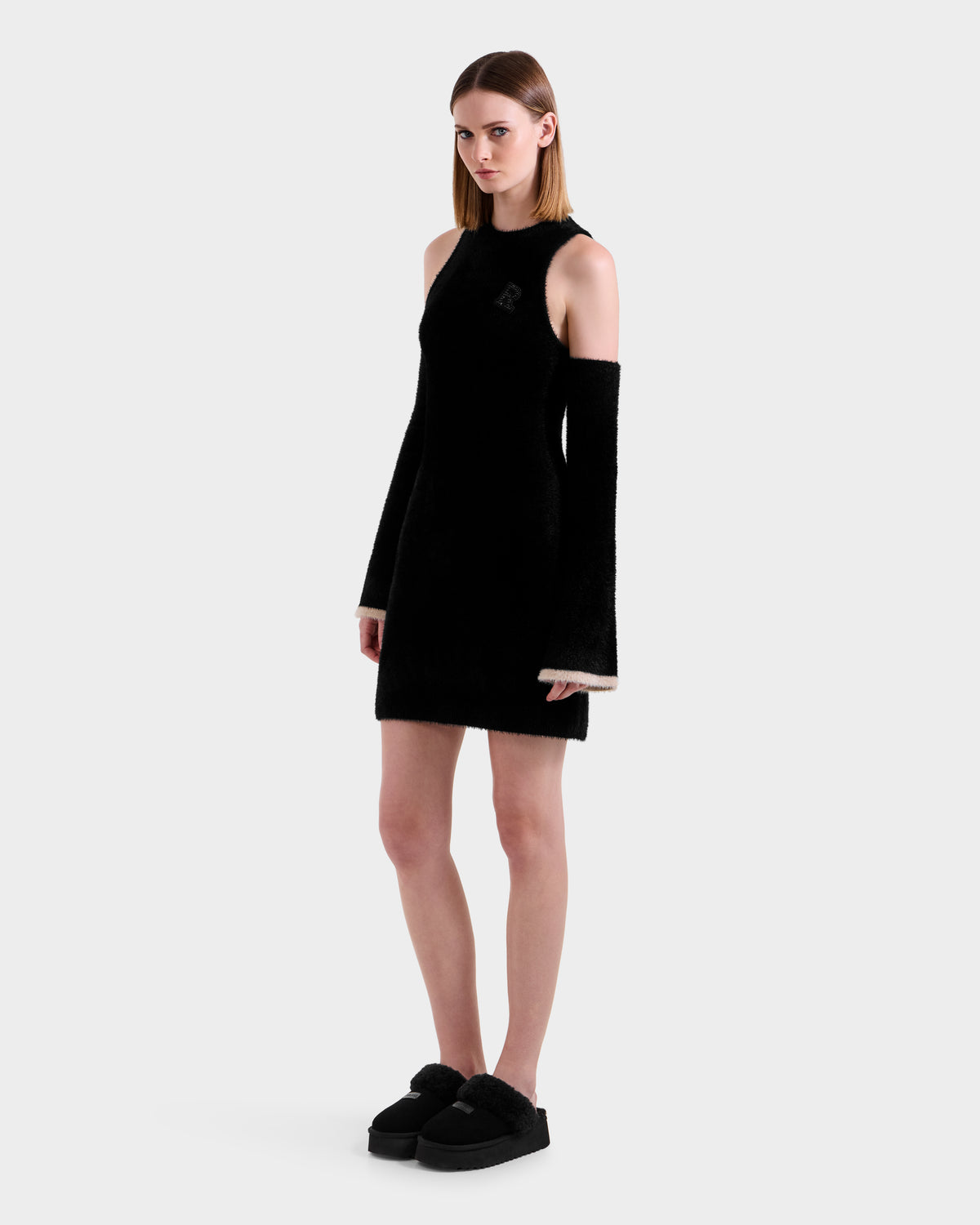 A woman stands against a plain white background wearing the Rudsak ZARAH - 8525696 Black long-sleeve cold-shoulder fitted dress and black platform slippers.