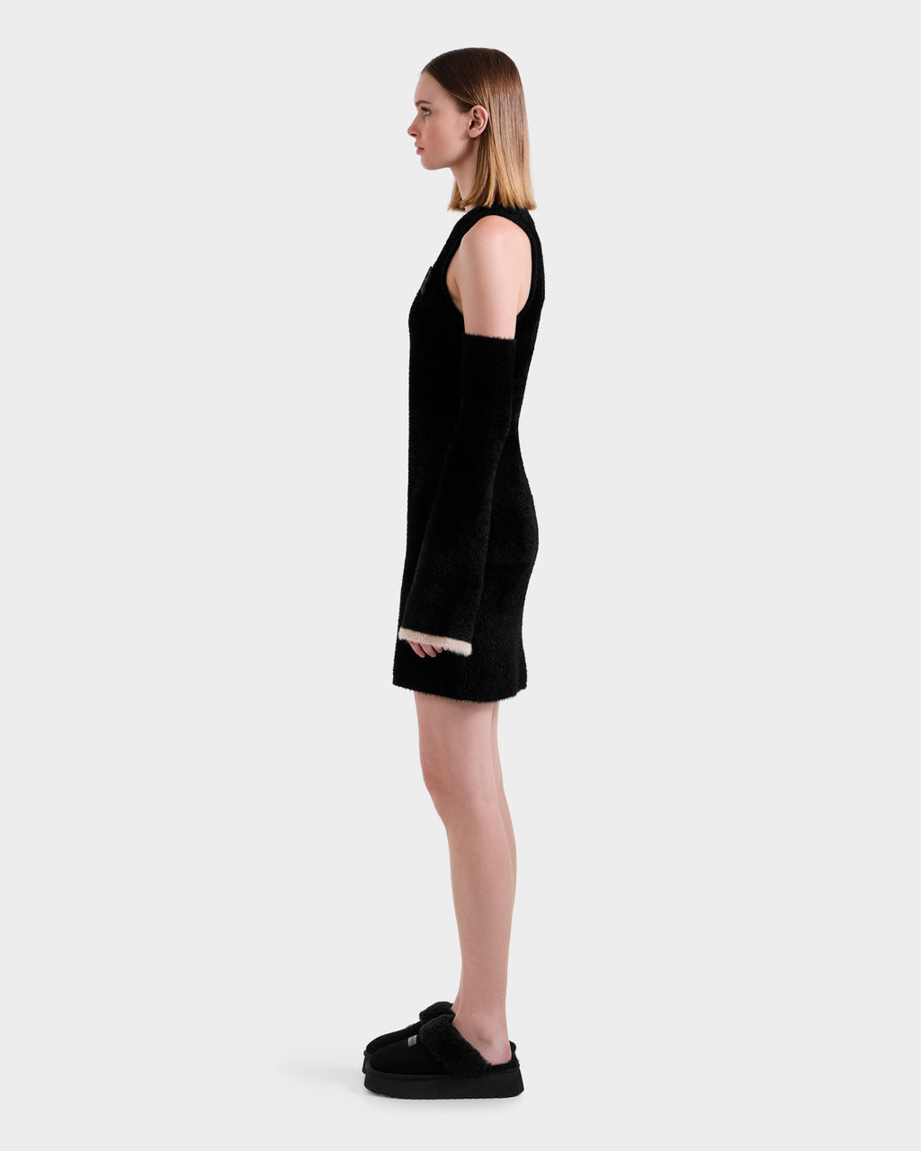 A woman in profile stands against a plain background, wearing the Rudsak ZARAH - 8525696 Black women's fitted dress with long detached sleeves and black platform shoes. Her straight, shoulder-length light brown hair completes the look.