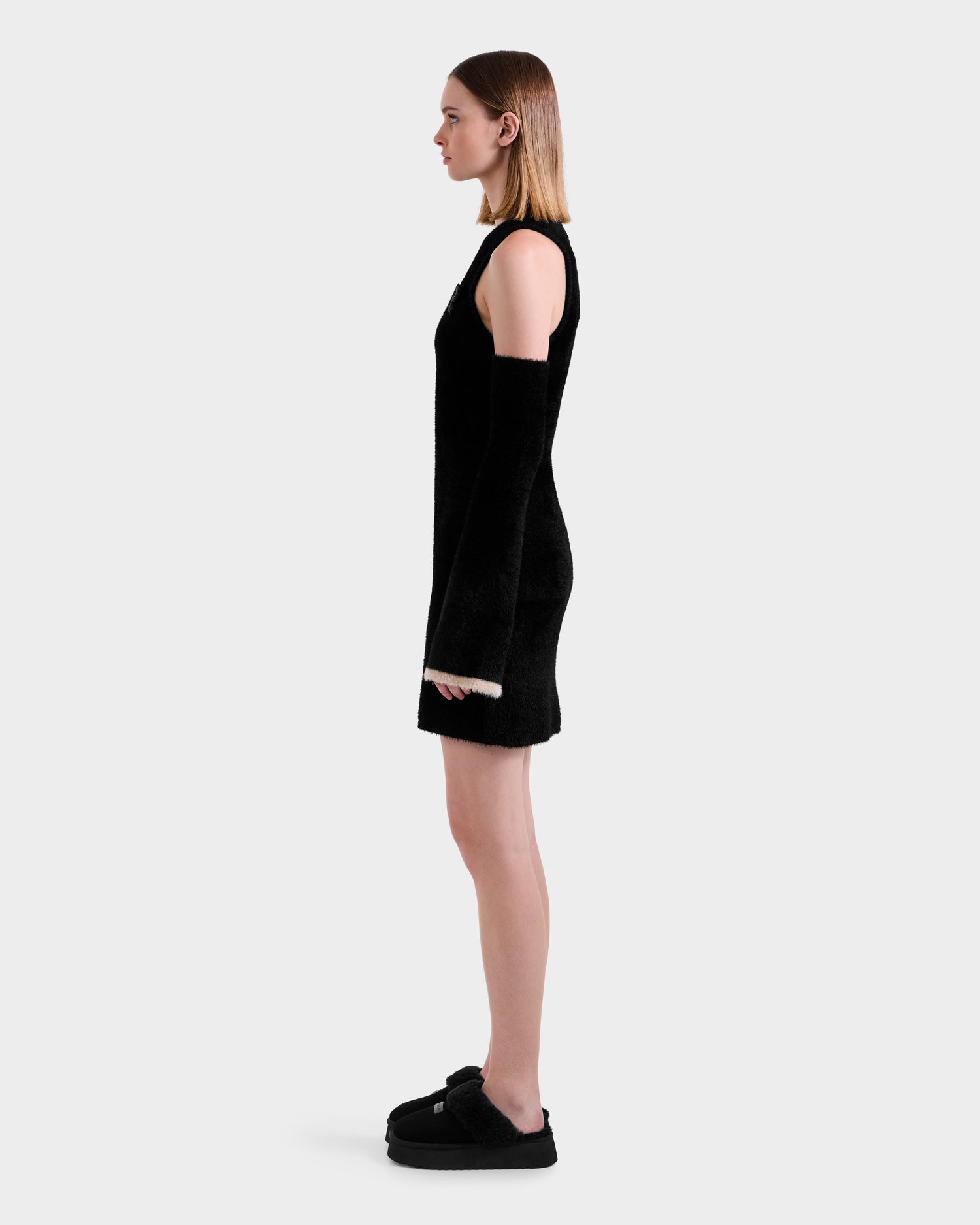 A woman in profile stands against a plain background, wearing the Rudsak ZARAH - 8525696 Black women's fitted dress with long detached sleeves and black platform shoes. Her straight, shoulder-length light brown hair completes the look.